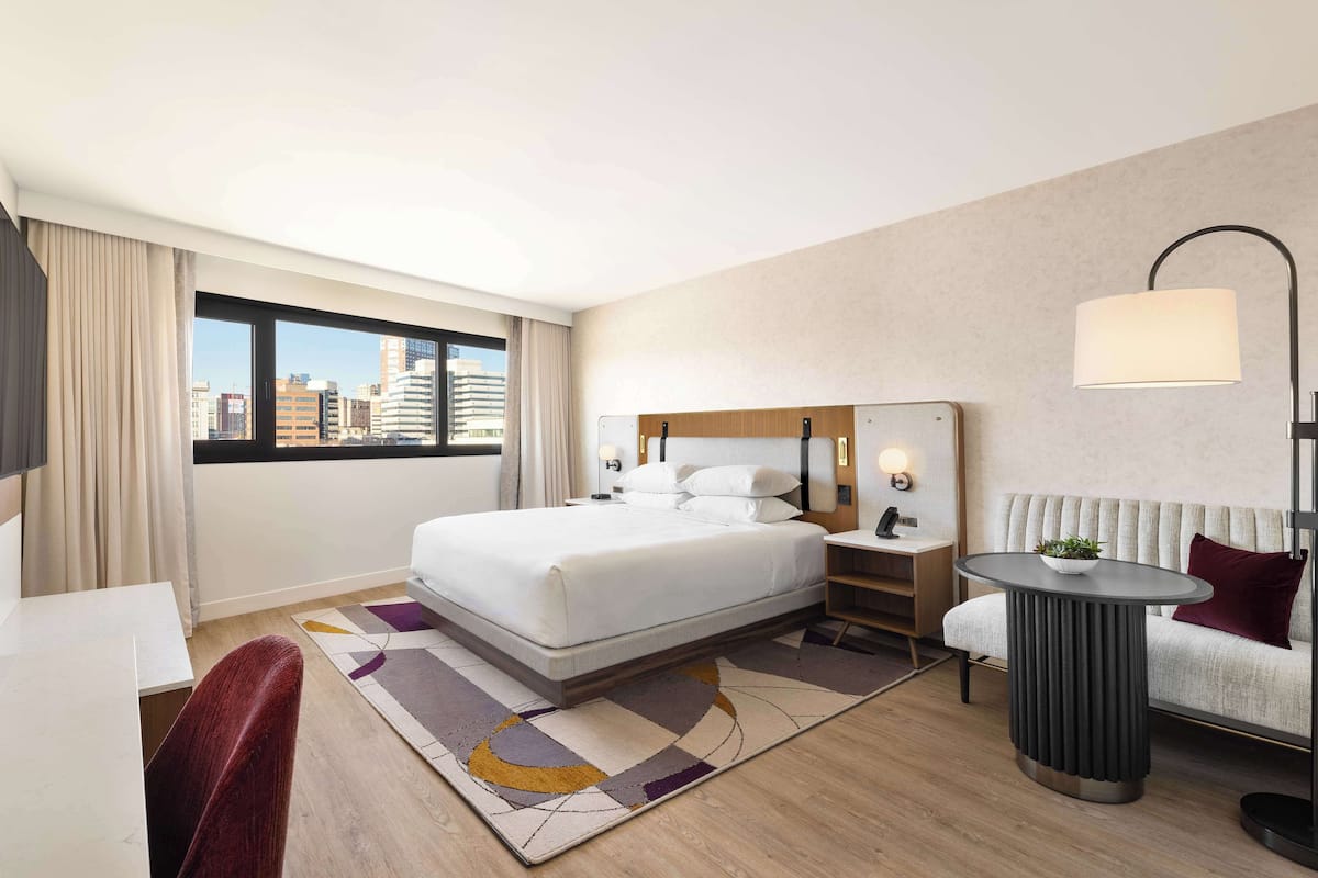 A modern hotel room with a large bed, a desk, a chair, a sofa, a lamp, and a view of the city.