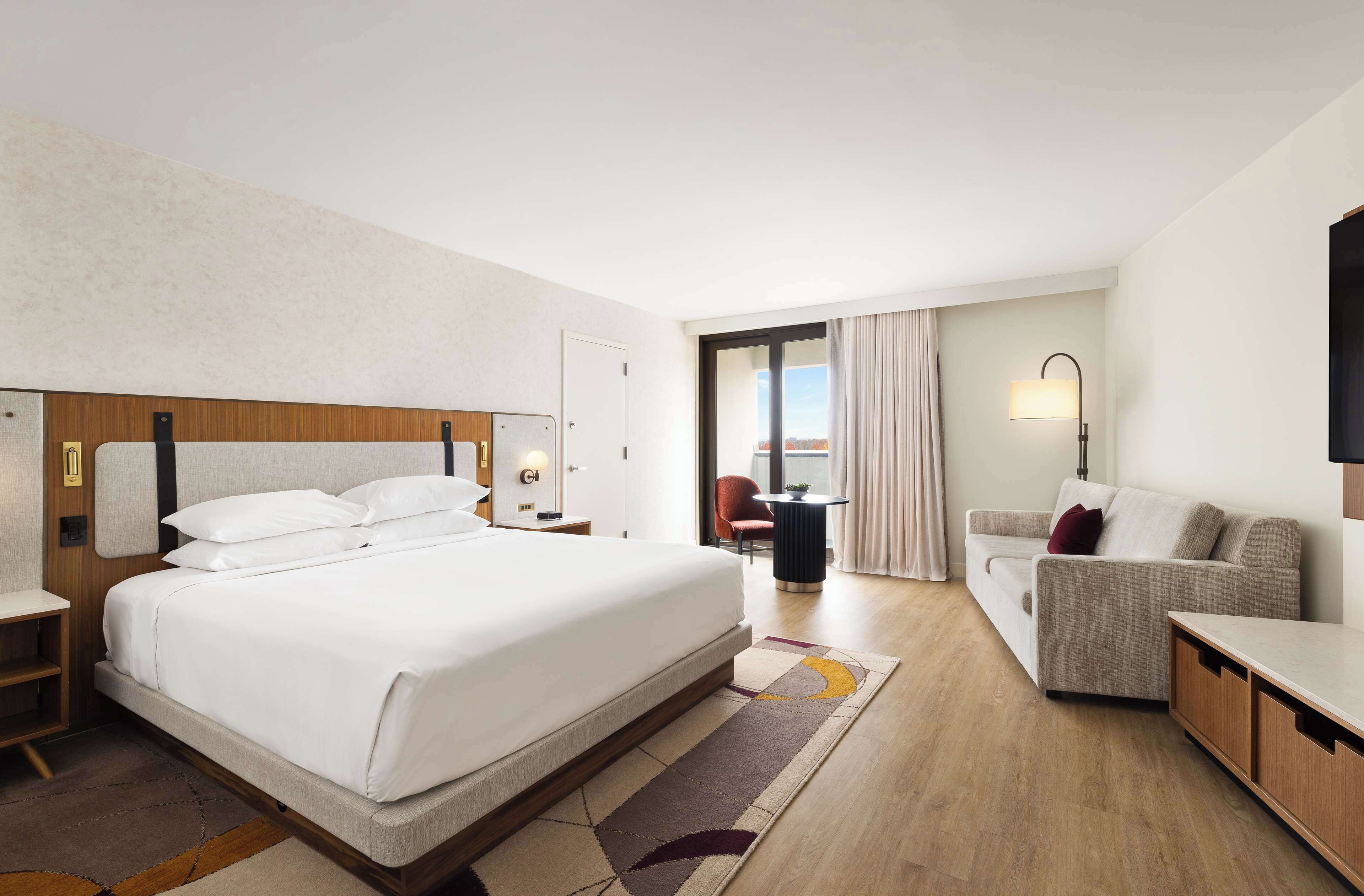 Quarto de hotel com uma cama grande, um sofá, uma escrivaninha e uma cadeira. Há uma janela com cortinas e vista para o exterior.