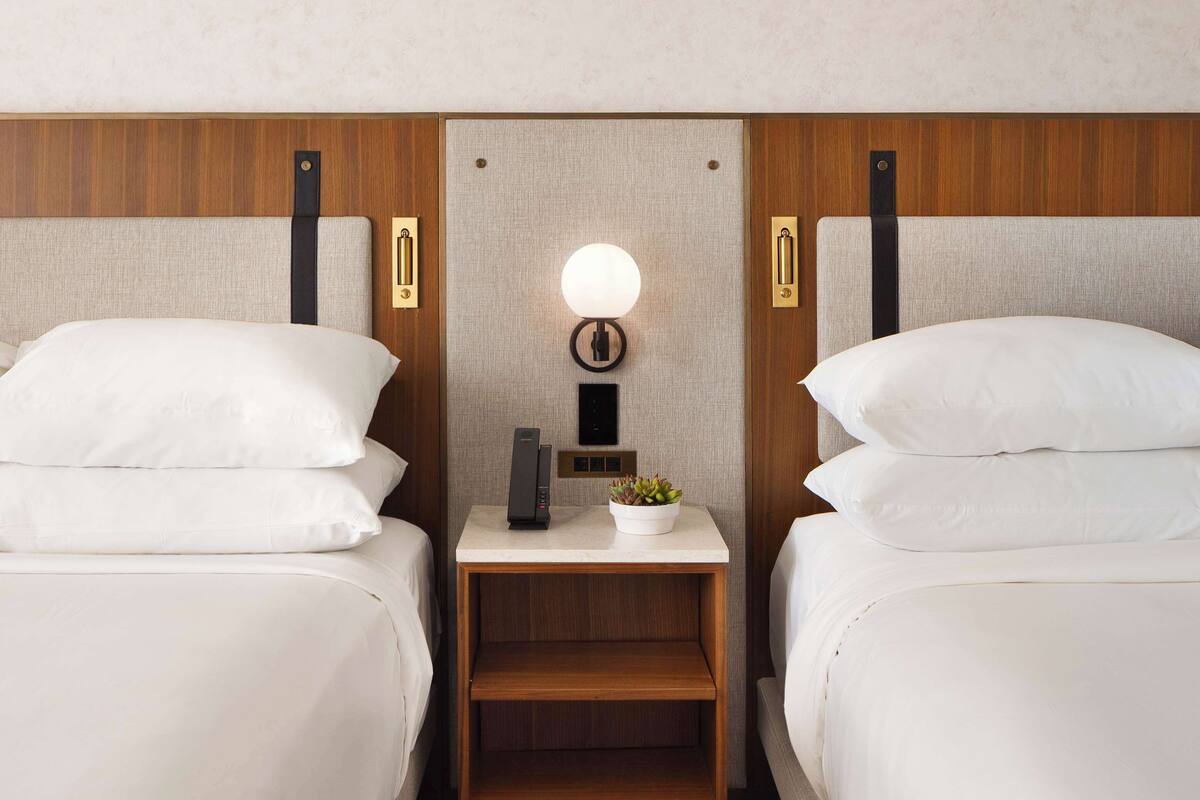 A hotel room with two single beds, a nightstand with a lamp, a small table, and a wall-mounted shelf.
