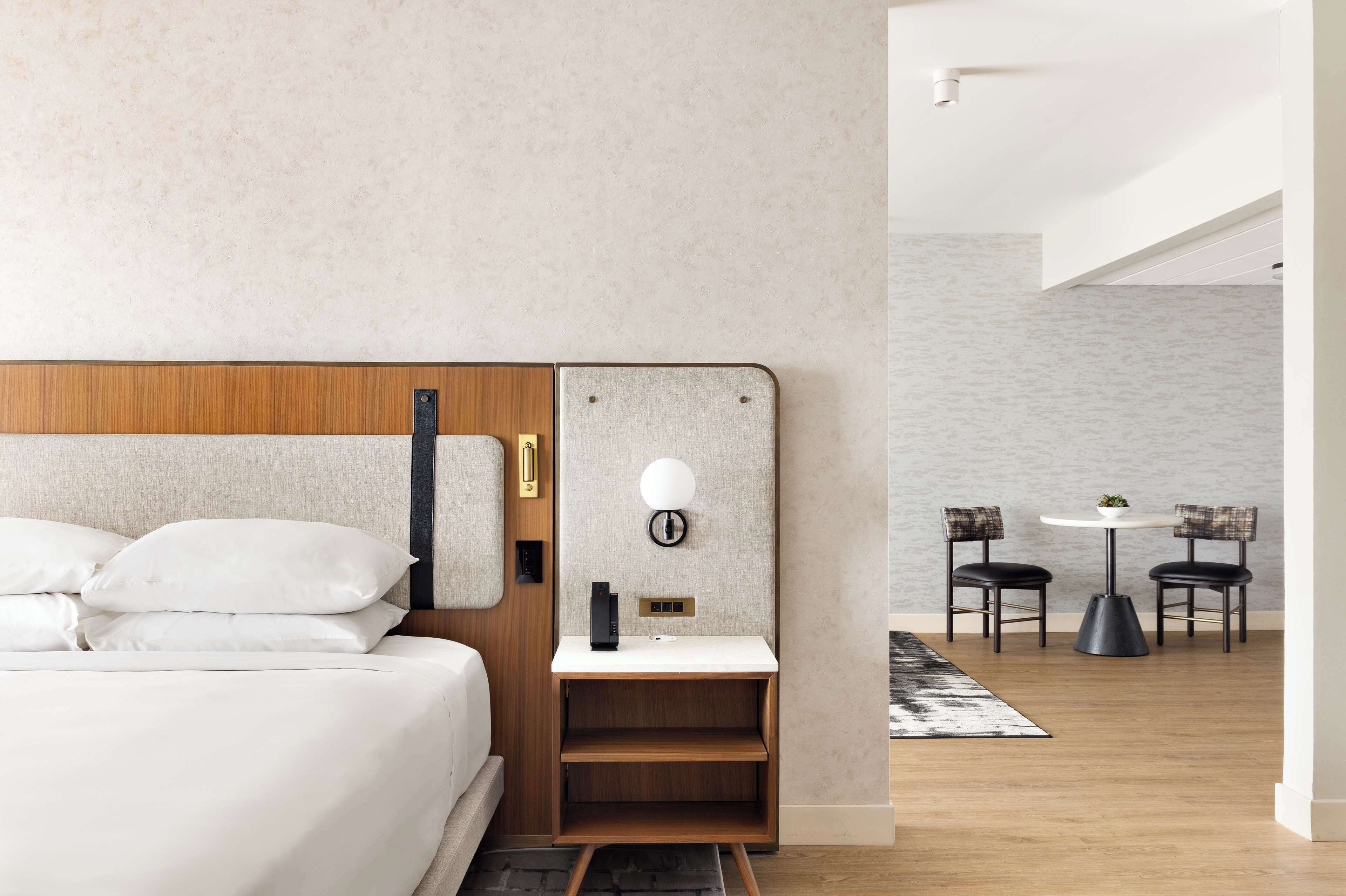 A modern hotel room with a wooden headboard, a bedside table with a lamp, and a small seating area with two chairs and a table.