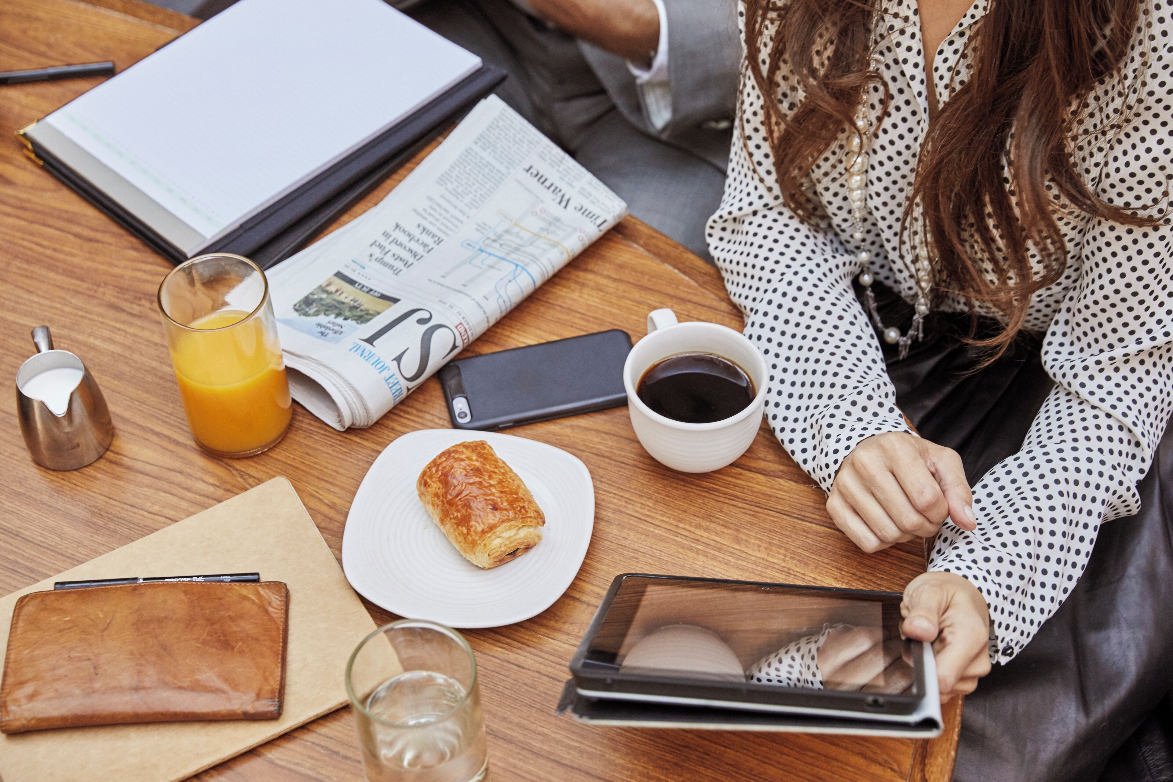 Une table en bois avec un journal, un verre de jus d’orange, une tasse de café, une pâtisserie, un portefeuille, un verre d’eau et une tablette.