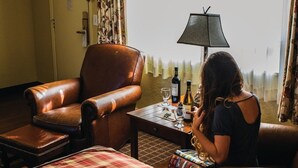 Een hotelkamer met een bruine leren fauteuil, een klein tafeltje met een fles en glazen, en een geruit beddegoed.