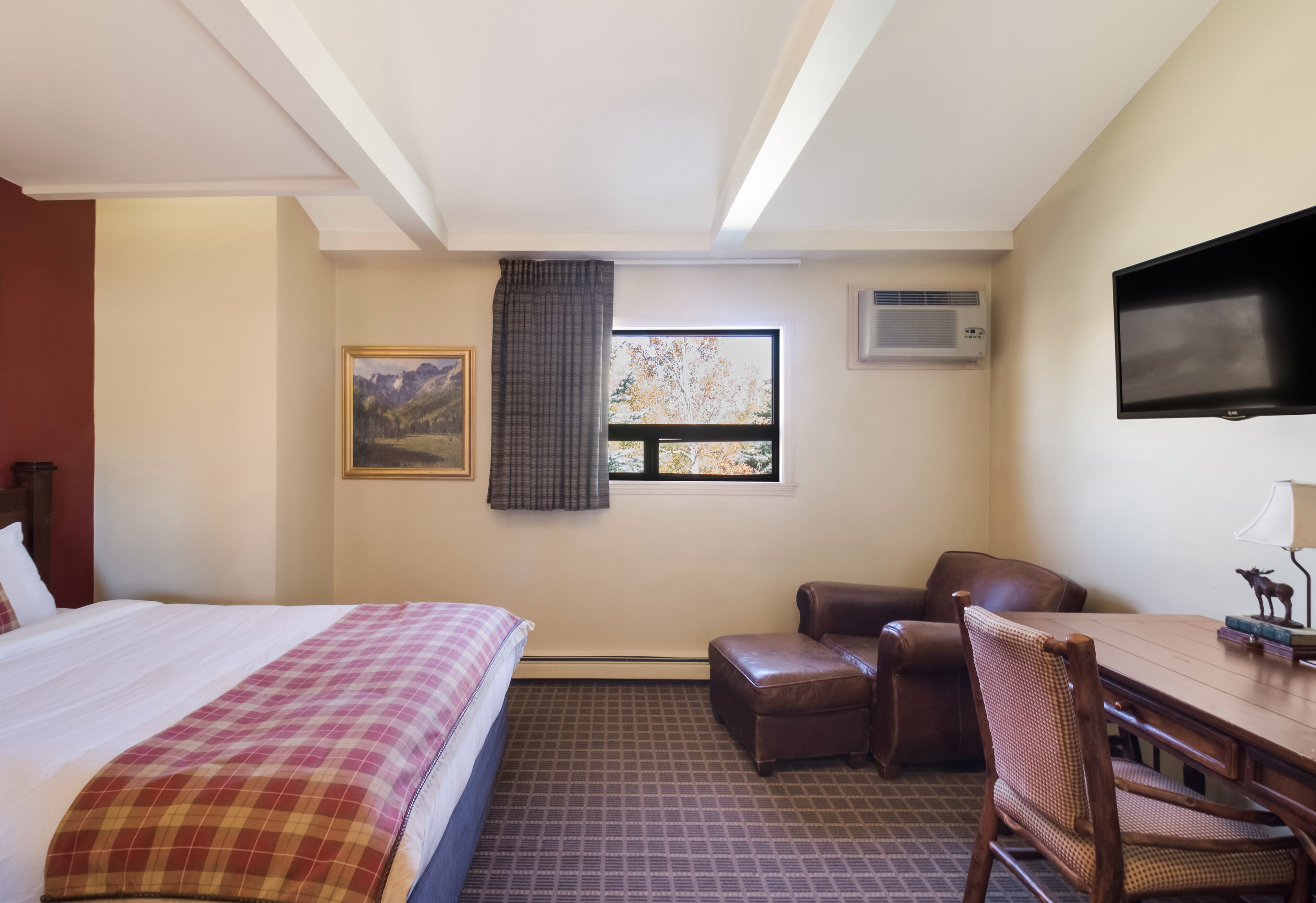 A hotel room with a bed, a desk, a chair, a television, and a window with a view of trees.