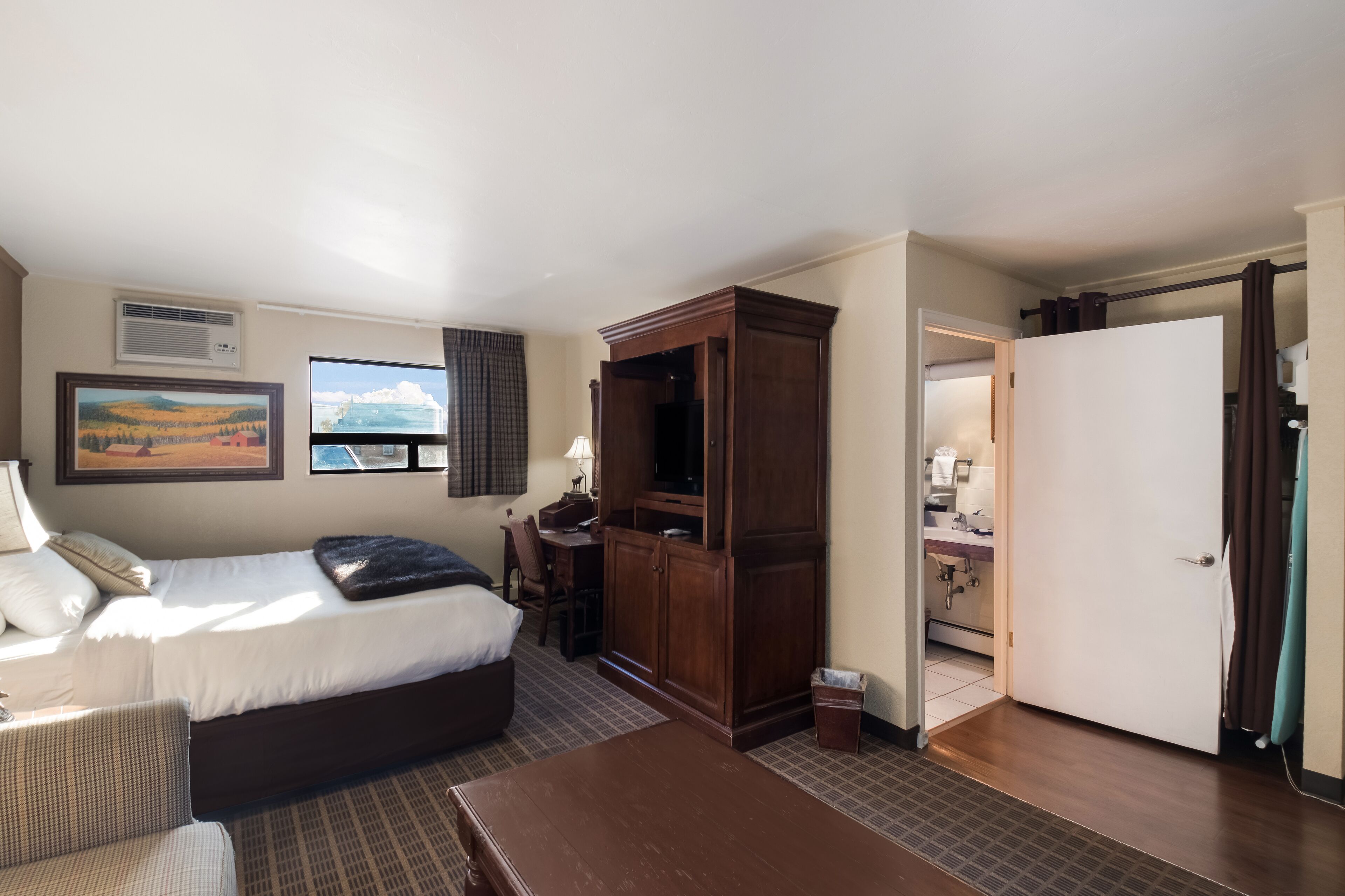 Quarto de hotel com cama, escrivaninha, guarda-roupa e casa de banho visível por uma porta aberta.