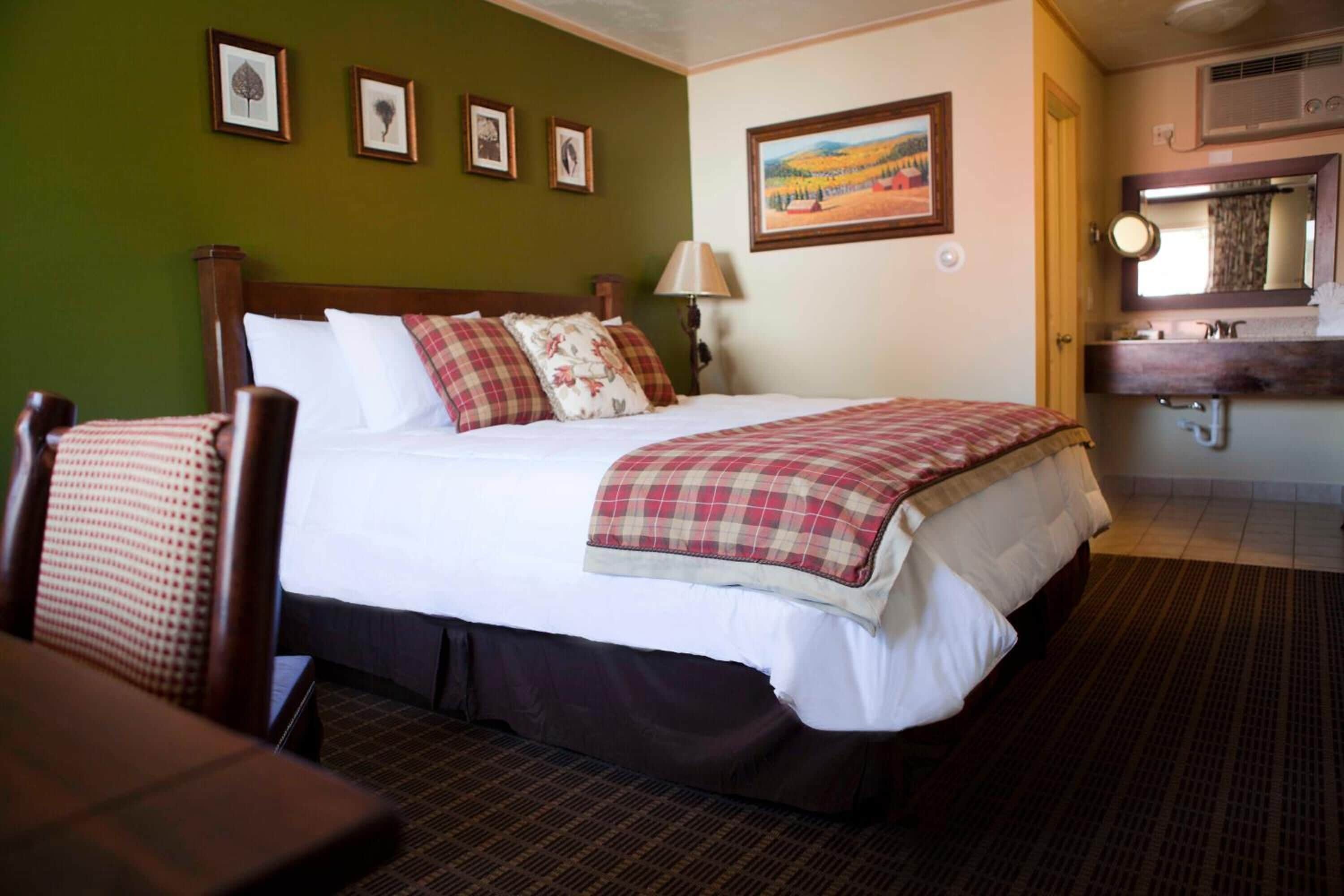 A hotel room with a bed, a chair, a desk, a painting on the wall, and a bathroom with a sink and mirror.