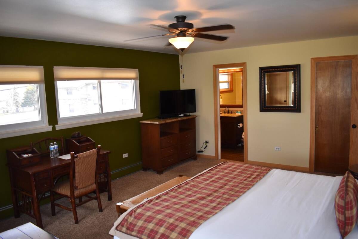 A bedroom with a bed, a desk with a chair, a television, and a ceiling fan.
