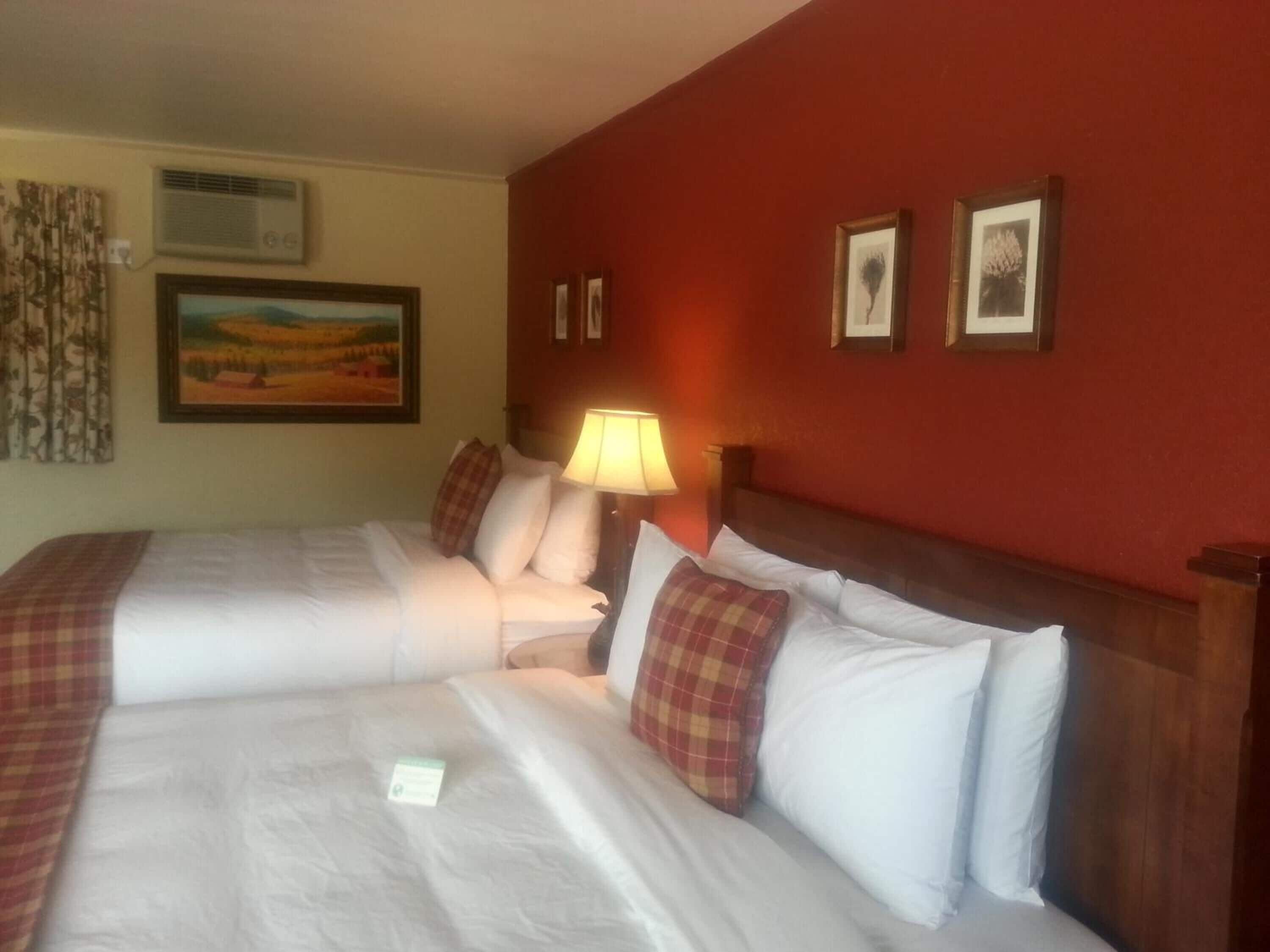 A hotel room with two beds, a nightstand with a lamp, a painting on the wall, and a window with curtains.