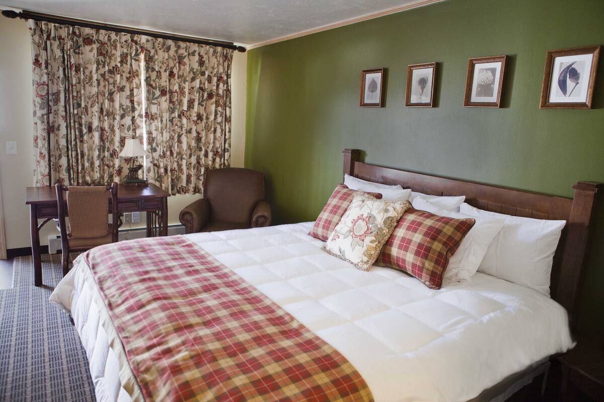 A hotel room with a large bed, a desk with a chair, a wooden nightstand, and a green wall with framed pictures.