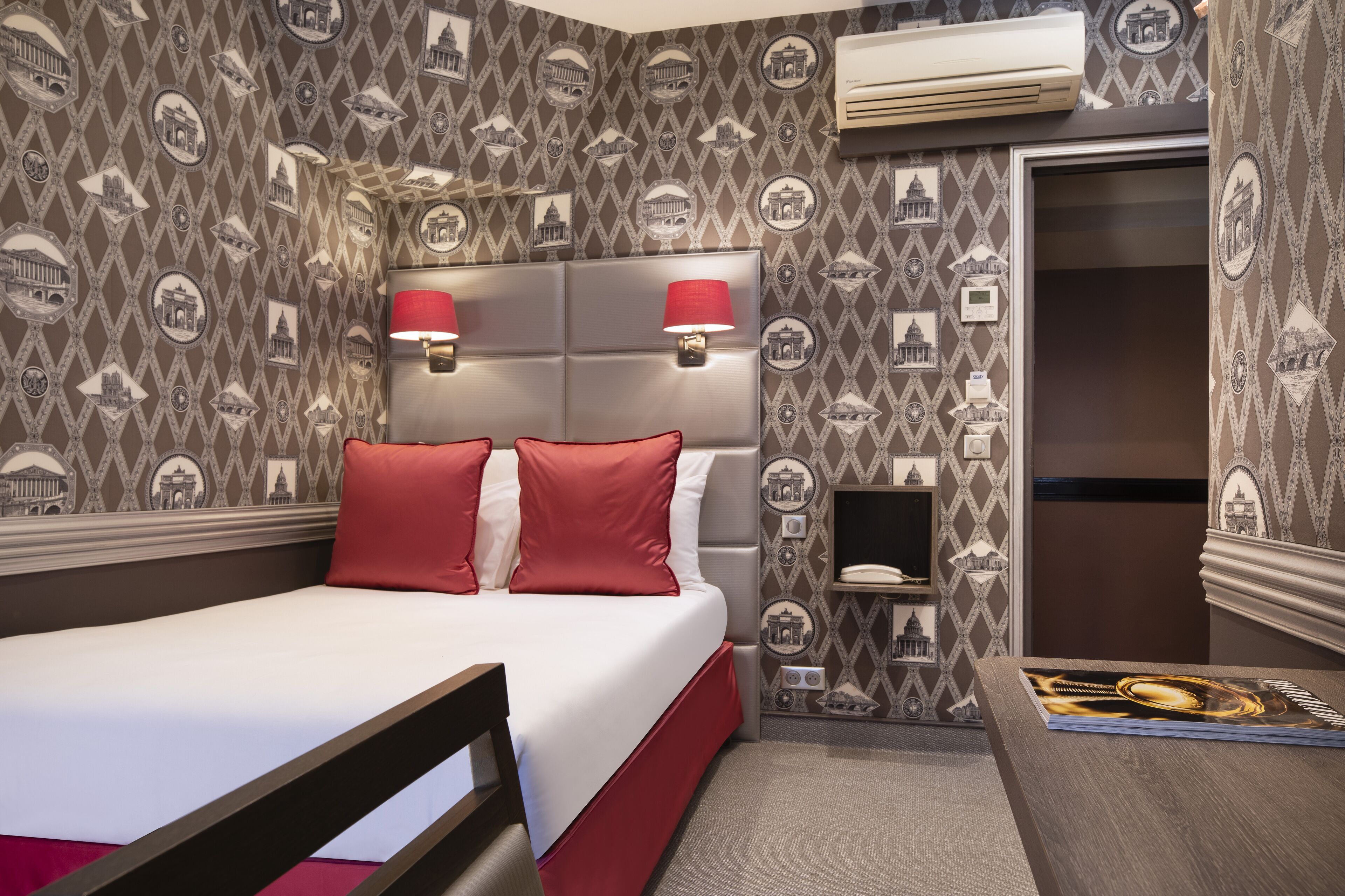 A hotel room with a bed, red pillows, a headboard, wall-mounted lamps, a bedside table, and a wall with a patterned design.