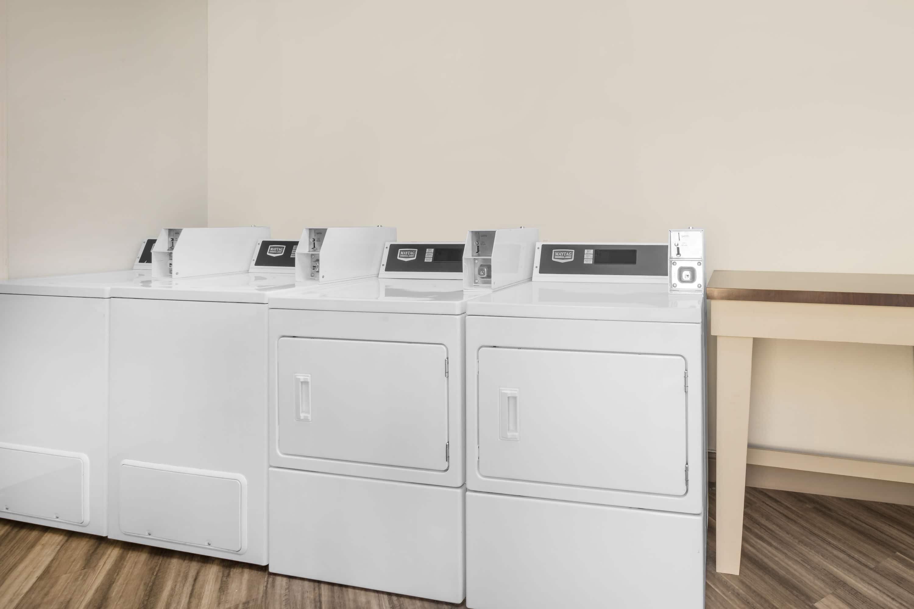 A row of white stacked washer and dryer units in a laundry room with a wooden floor and a light-colored wall.