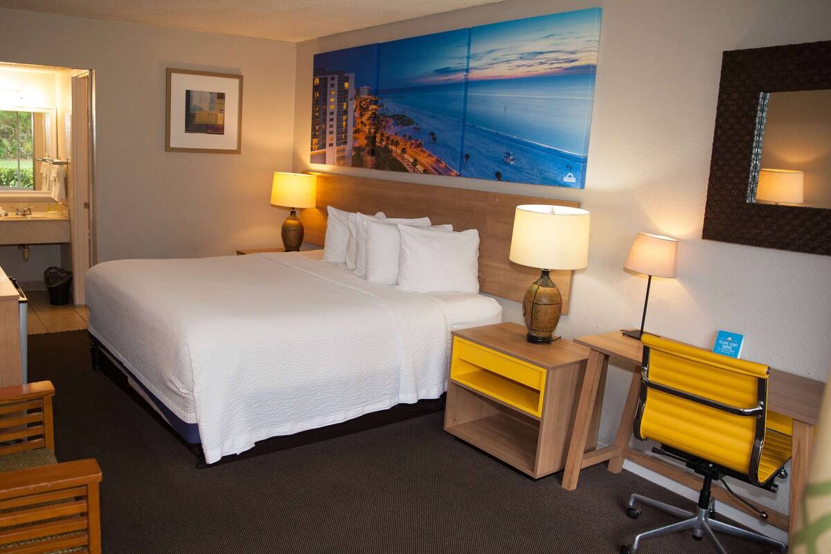 Een hotelkamer met een groot bed, twee nachtkastjes met lampen, een bureau met een stoel, een schilderij van een strandlandschap en een spiegel aan de muur.