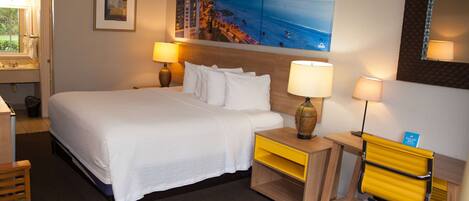 Una habitación de hotel con una cama grande, dos mesitas de noche con lámparas, un escritorio con una silla, un cuadro de una playa y un espejo en la pared.