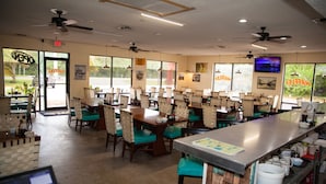Un restaurante con varias mesas y sillas, ventiladores de techo y grandes ventanales.