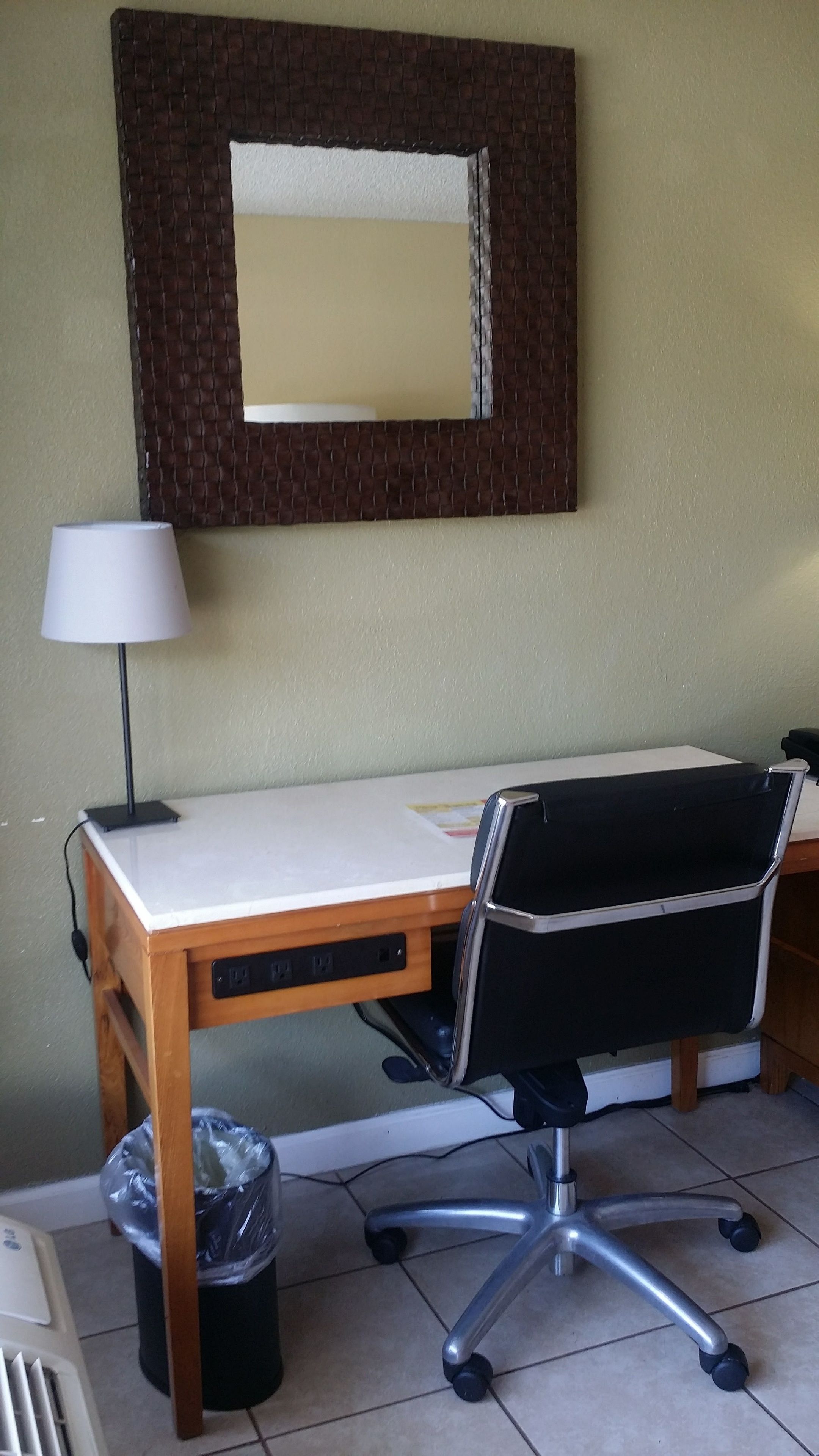 A hotel room with a wooden desk, a black office chair, a white lamp, and a trash bin.