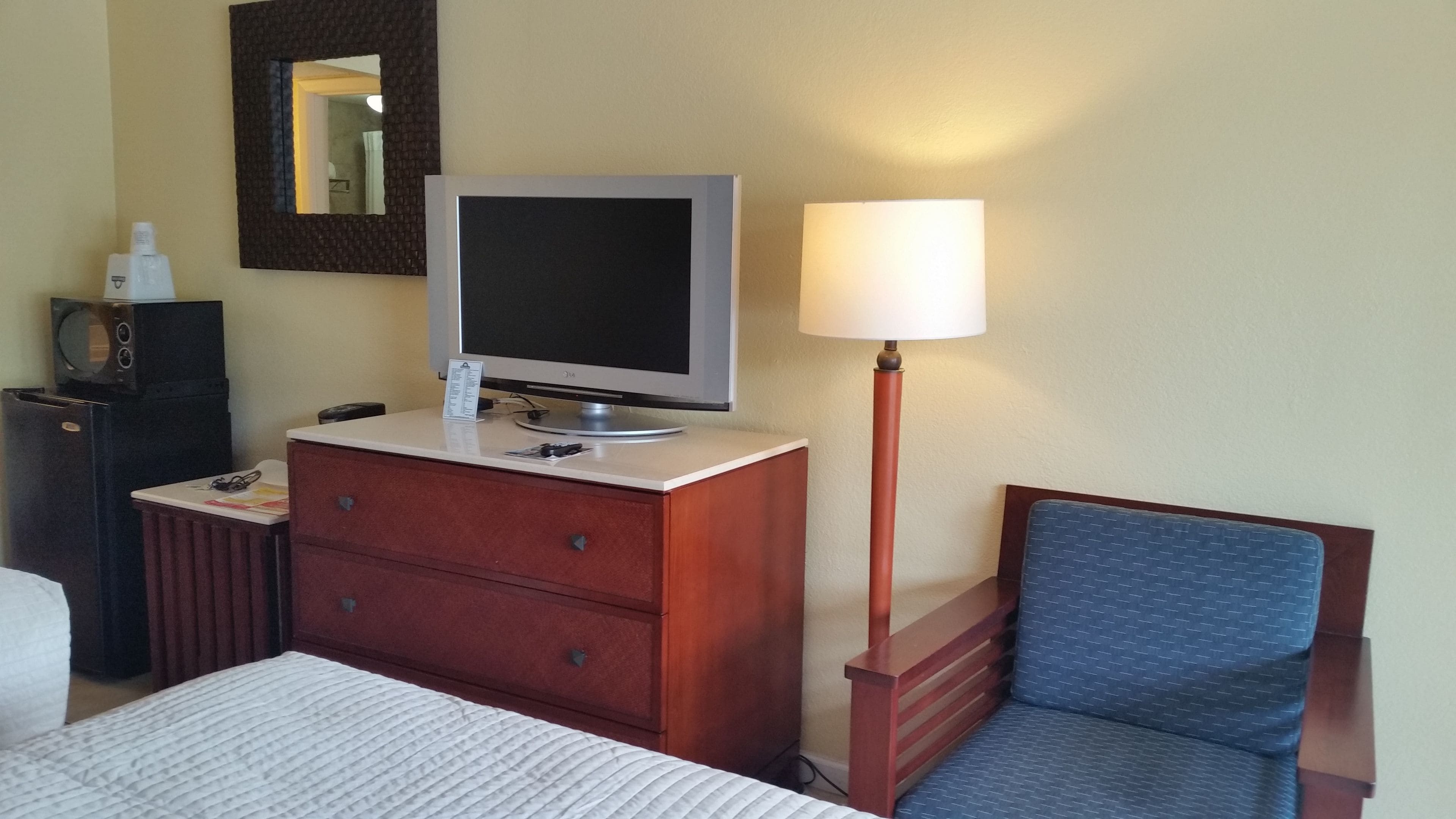 Una camera d'albergo con un letto, un comò in legno con la televisione, una lampada e una sedia.