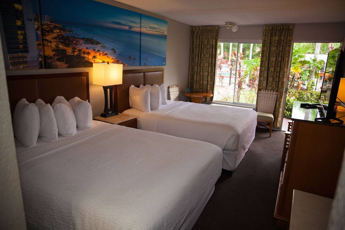 Een hotelkamer met twee bedden, een nachtkastje met een lamp, een bureau en uitzicht op een tuin door het raam.