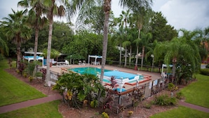 Zona de piscina rodeada de palmeras y vegetación, con sillas de descanso y un área de estar cubierta.