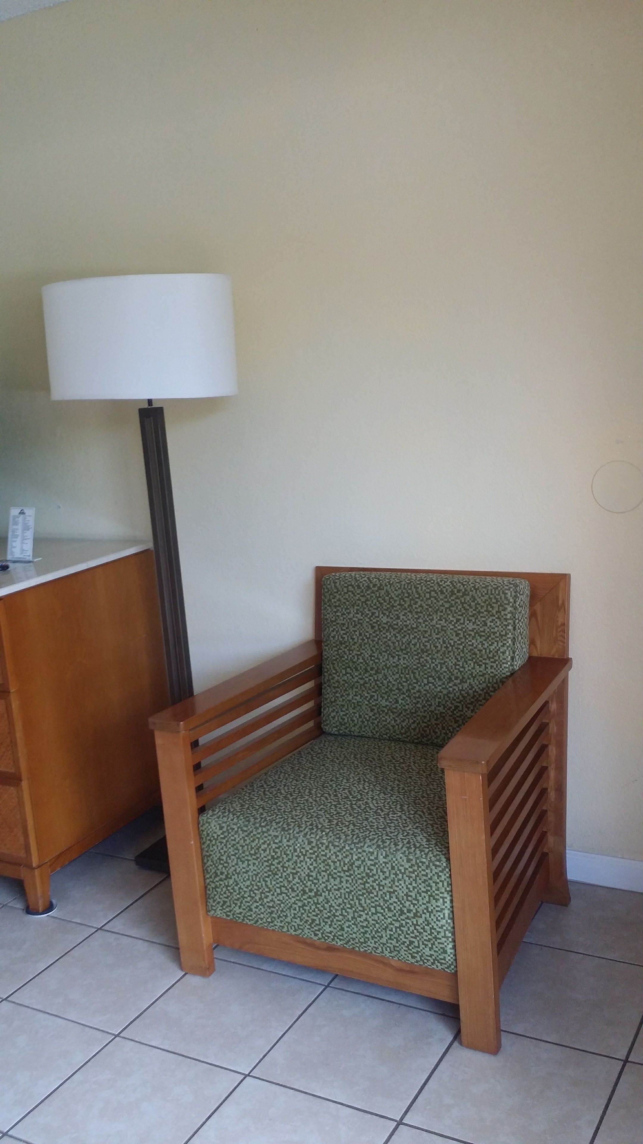 A corner of a room with a wooden chair with a patterned cushion, a desk with a lamp, and a tiled floor.