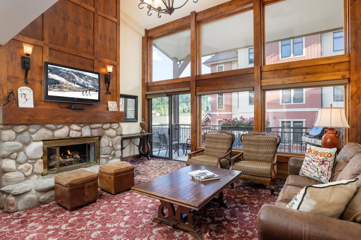 A cozy living room with a stone fireplace, wooden walls, and large windows offering a view of the outside.