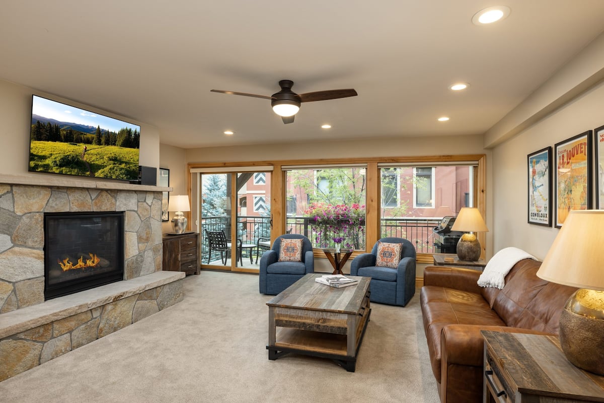 A living room with a stone fireplace, a flat-screen TV, a ceiling fan, and a large window with a view of a building and trees.
