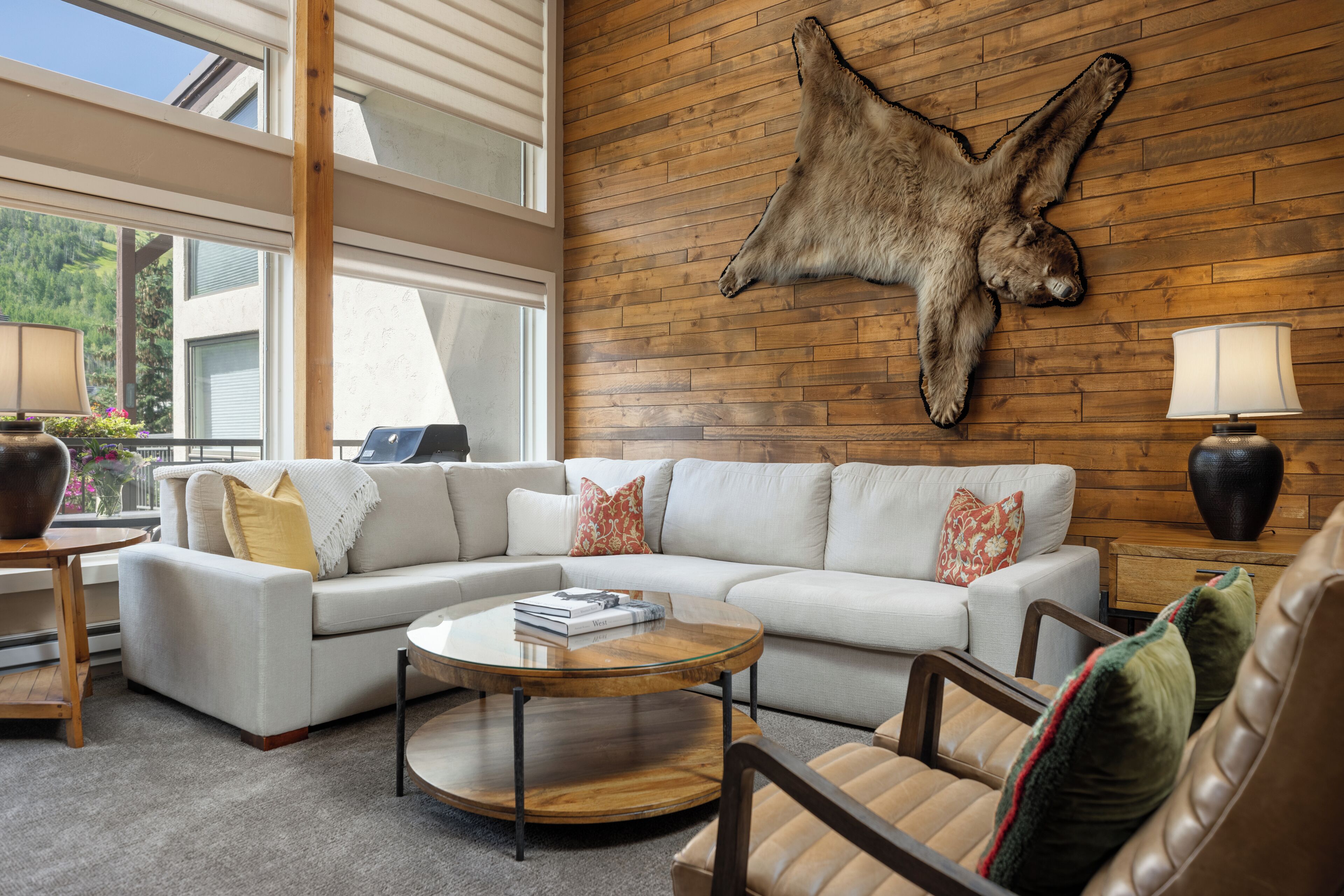 A modern living room with a sectional sofa, a round coffee table, leather armchairs, a wooden wall with a mounted animal hide, and a window with a view of greenery.