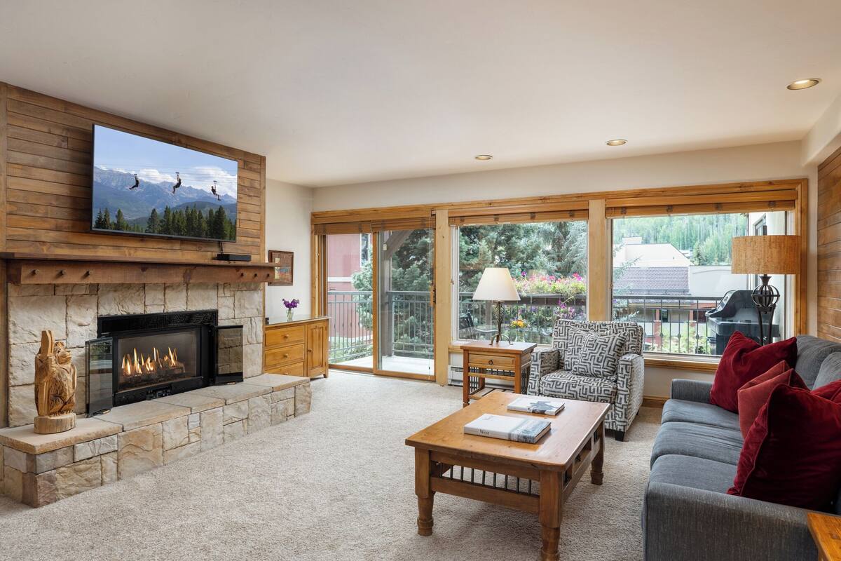 A living room with a fireplace, a television mounted above it, a sofa, a coffee table, and a view of the outdoors through large windows.