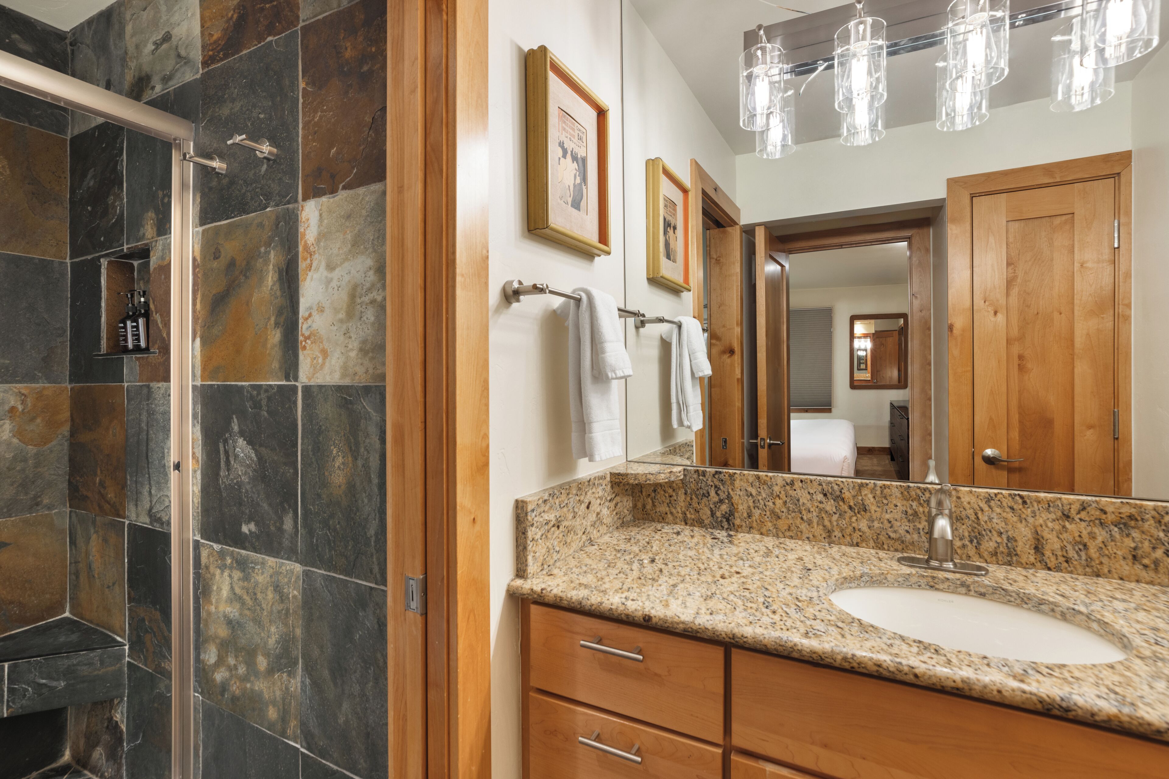 A bathroom with a granite countertop, a sink, a shower with stone tiles, wooden cabinets, and a mirror with framed pictures.