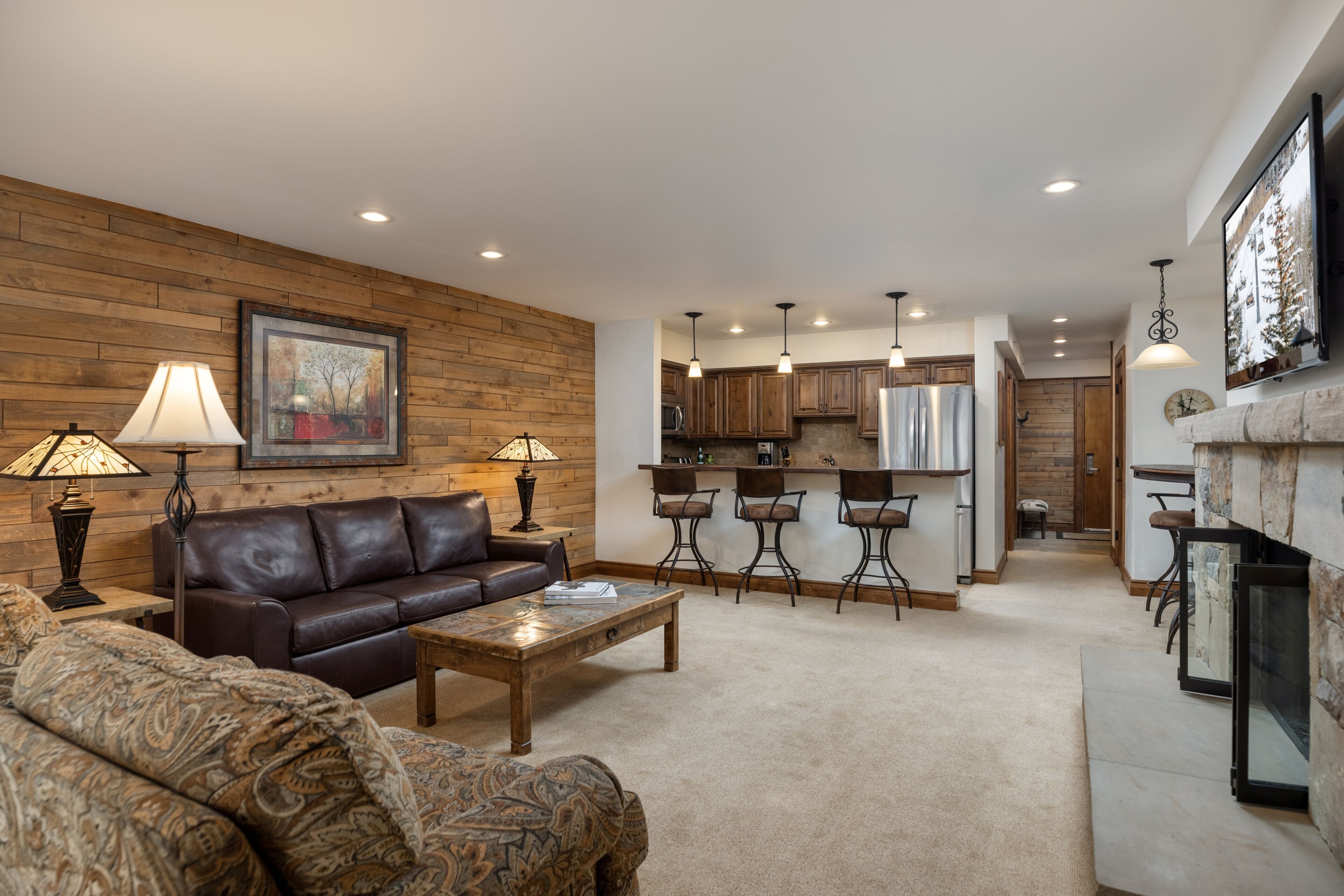 A spacious living room with a leather sofa, patterned armchair, and a coffee table. A kitchen with wooden cabinets and pendant lighting is visible in the background.