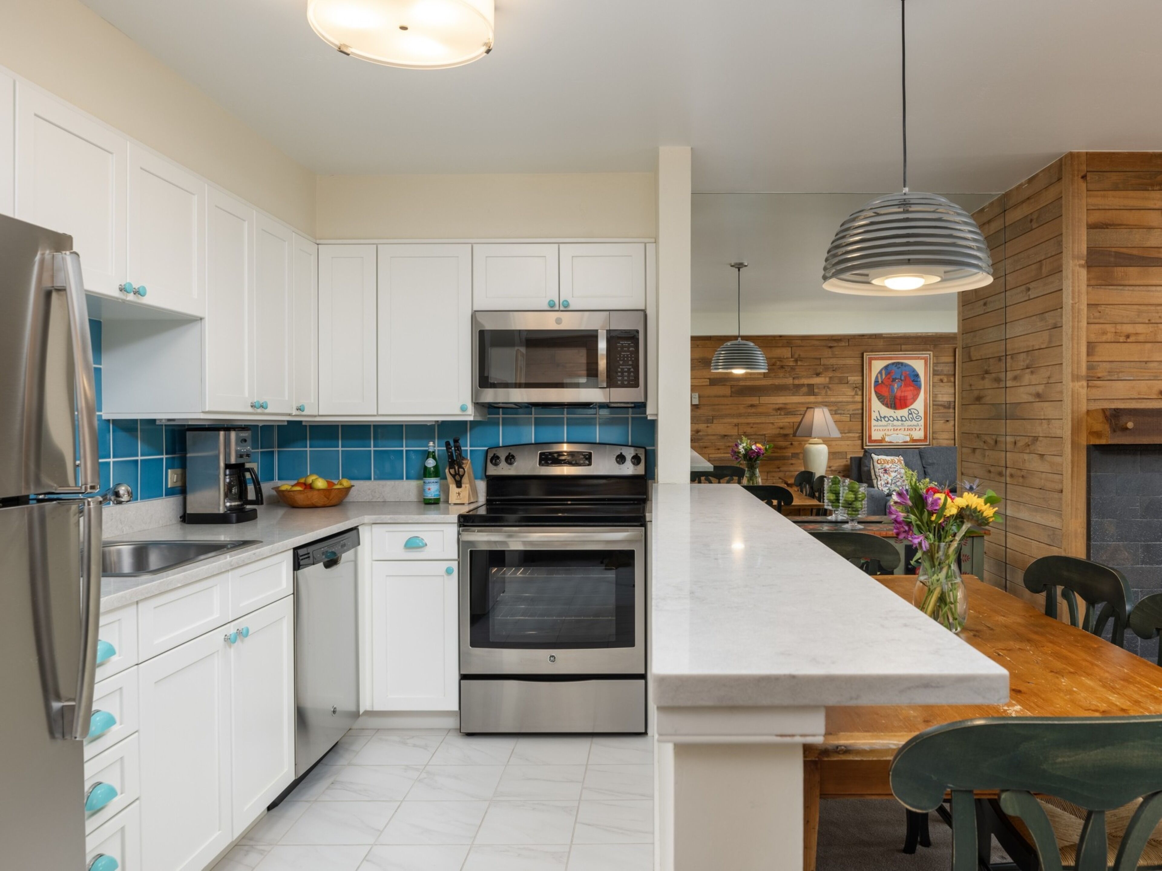 A modern kitchen with white cabinets, stainless steel appliances, a marble countertop, and a wooden dining area.