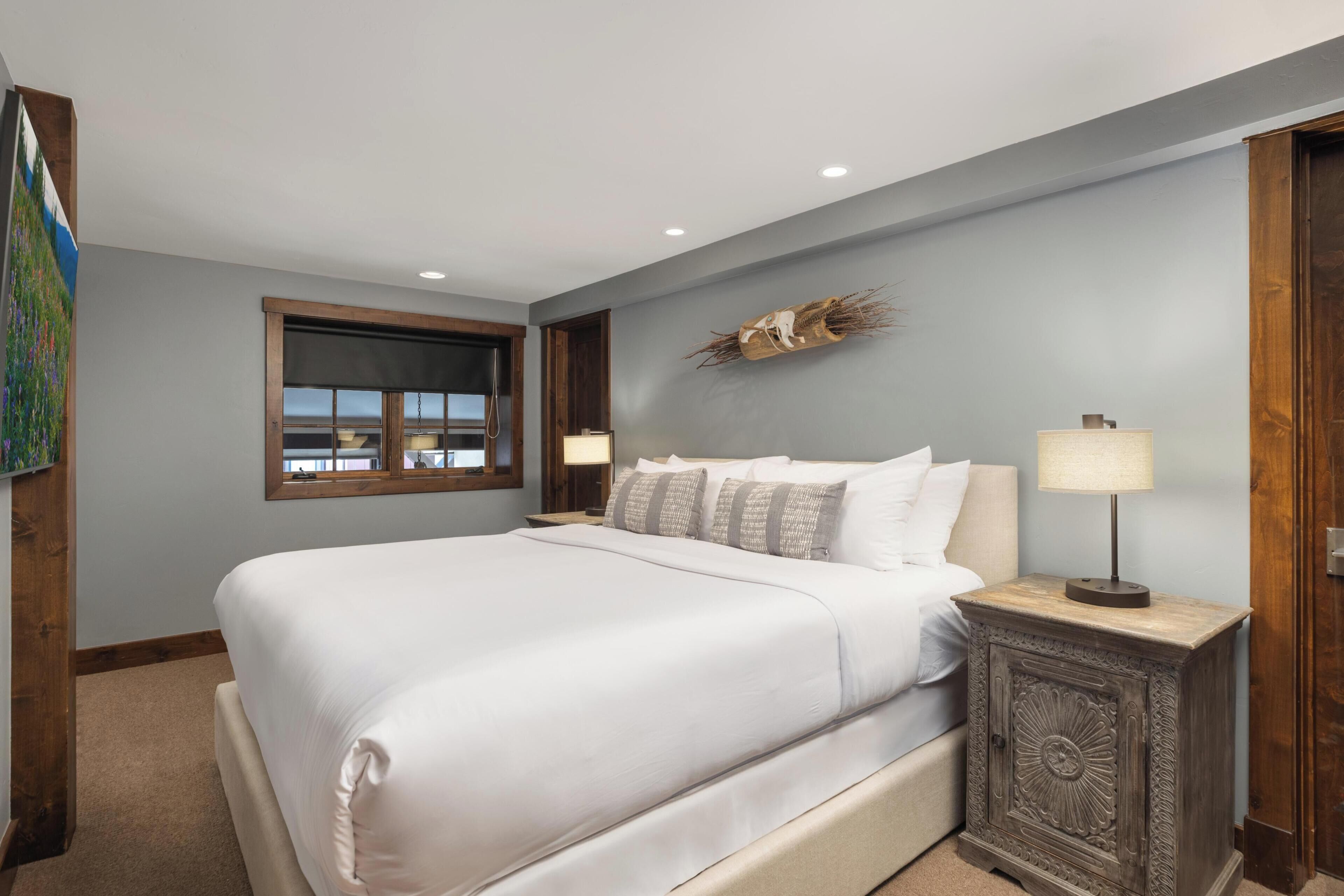 A bedroom with a large bed, a nightstand with a lamp, a window with blinds, and a wall-mounted decorative piece.