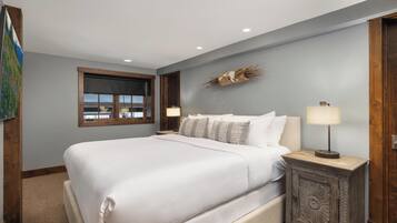 A bedroom with a large bed, a nightstand with a lamp, a window with blinds, and a wall-mounted decorative piece.