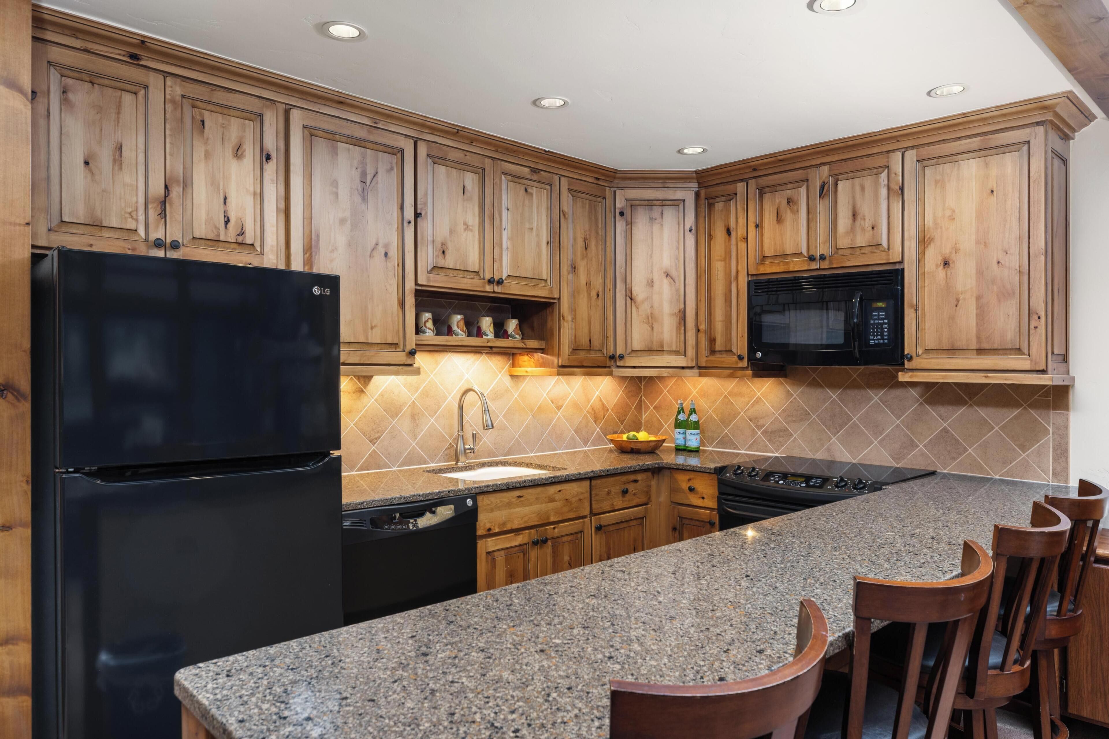 A kitchen with wooden cabinets, a black refrigerator, a microwave, a granite countertop, and bar stools.