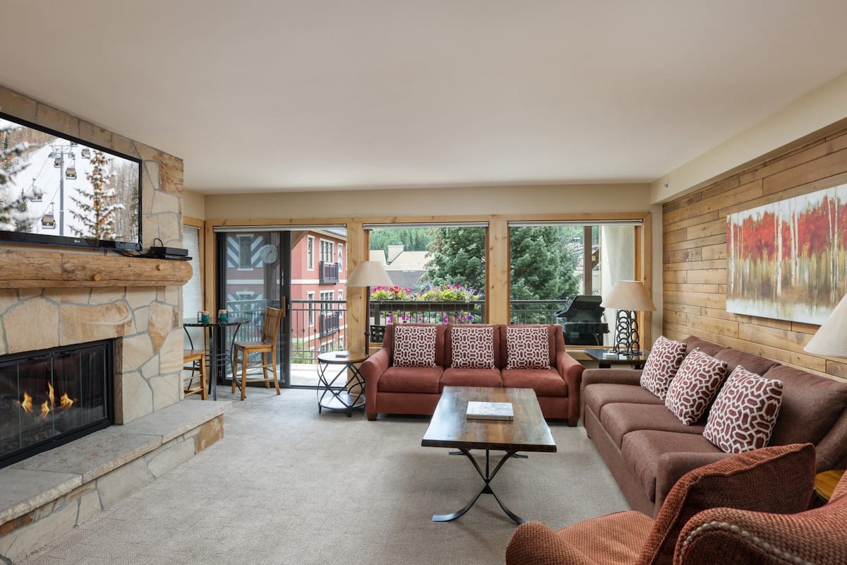 A cozy living room with a stone fireplace, a sofa, a coffee table, and a view of the outside through large windows.