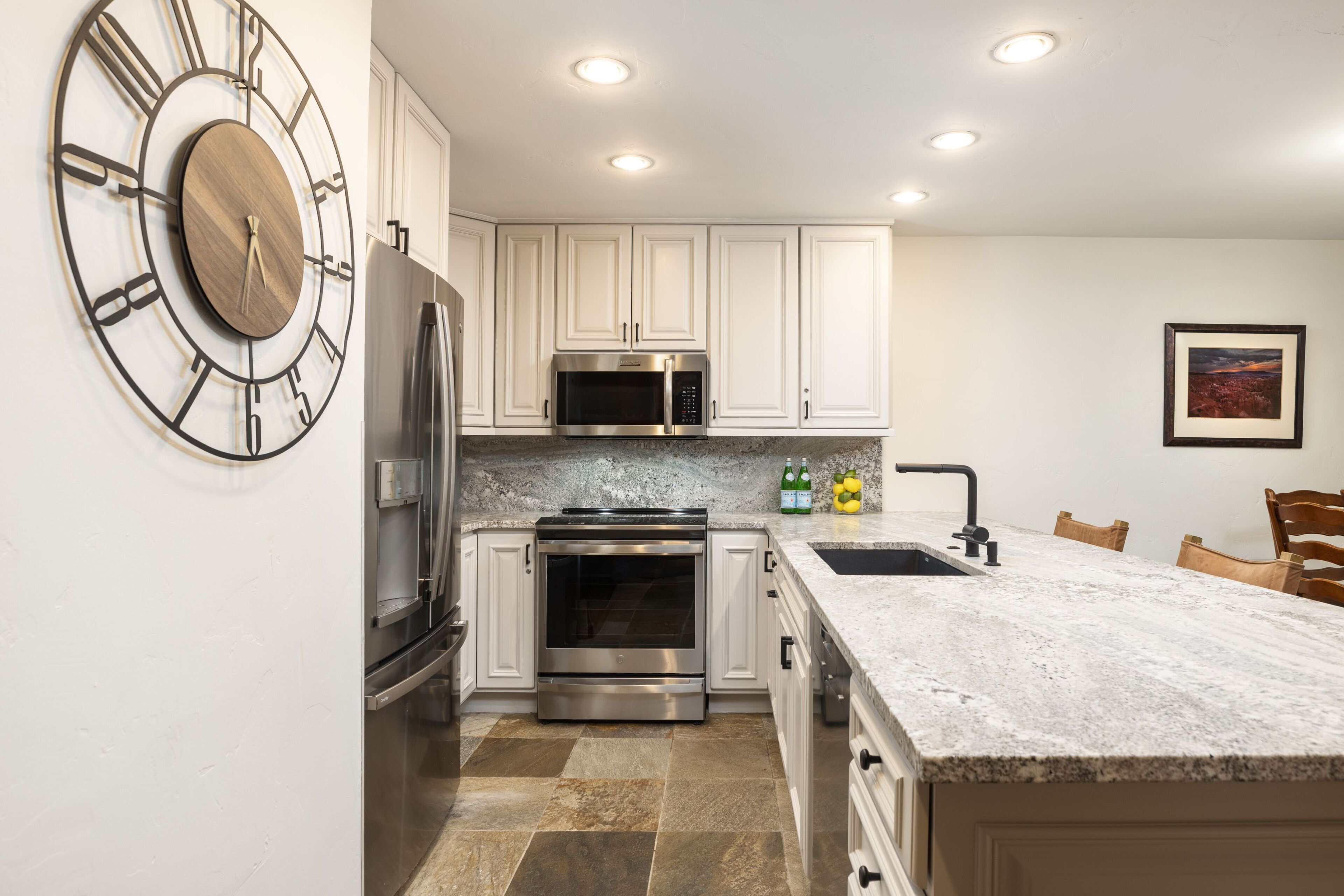 A modern kitchen with white cabinets, stainless steel appliances, a granite countertop, and a wall-mounted clock.