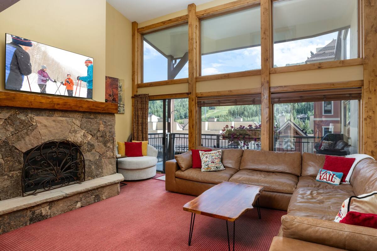 A cozy living room with a stone fireplace, a leather sofa, and a flat-screen TV displaying a family skiing.