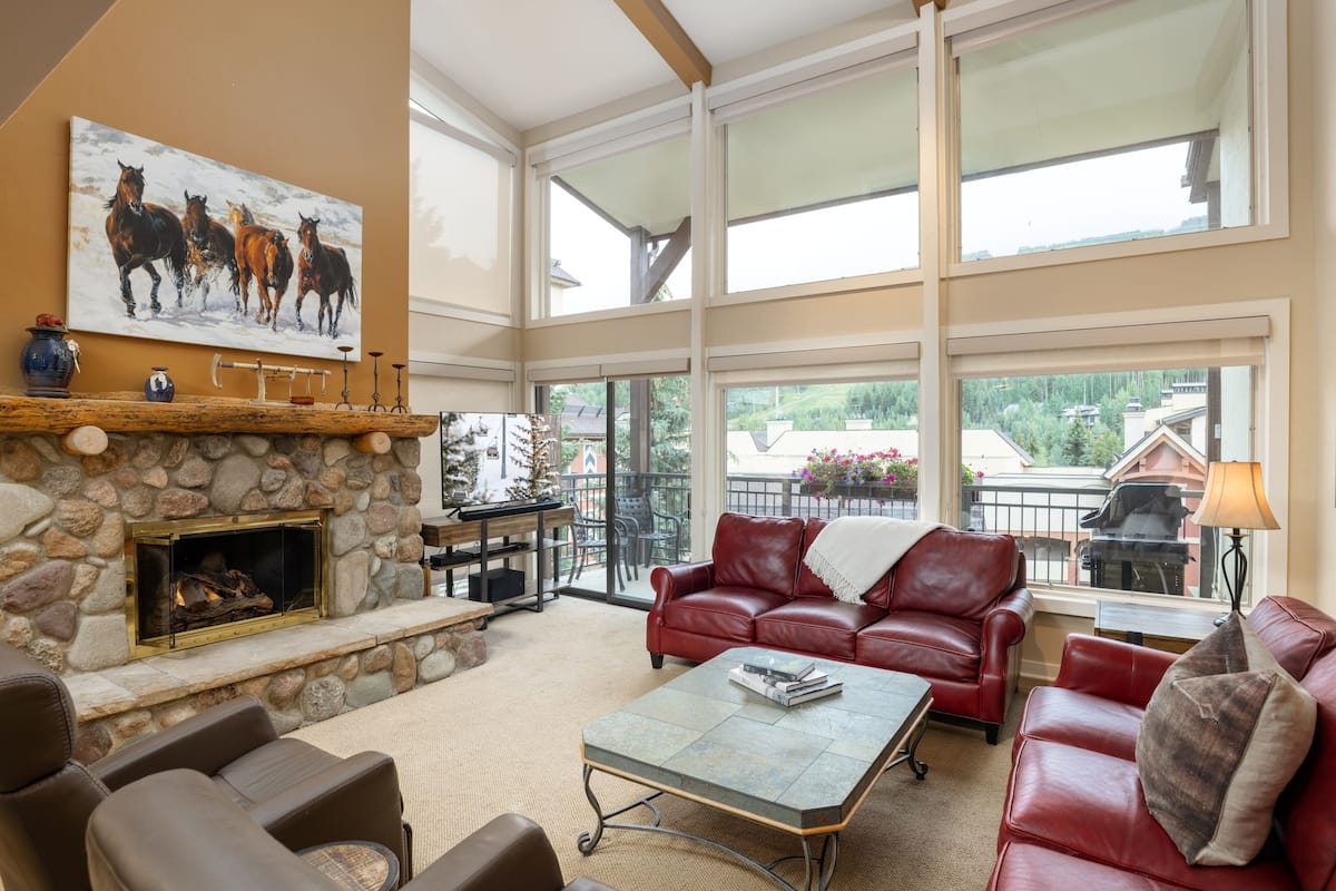 A spacious living area with a stone fireplace, leather furniture, and large windows offering a view of the outdoors.