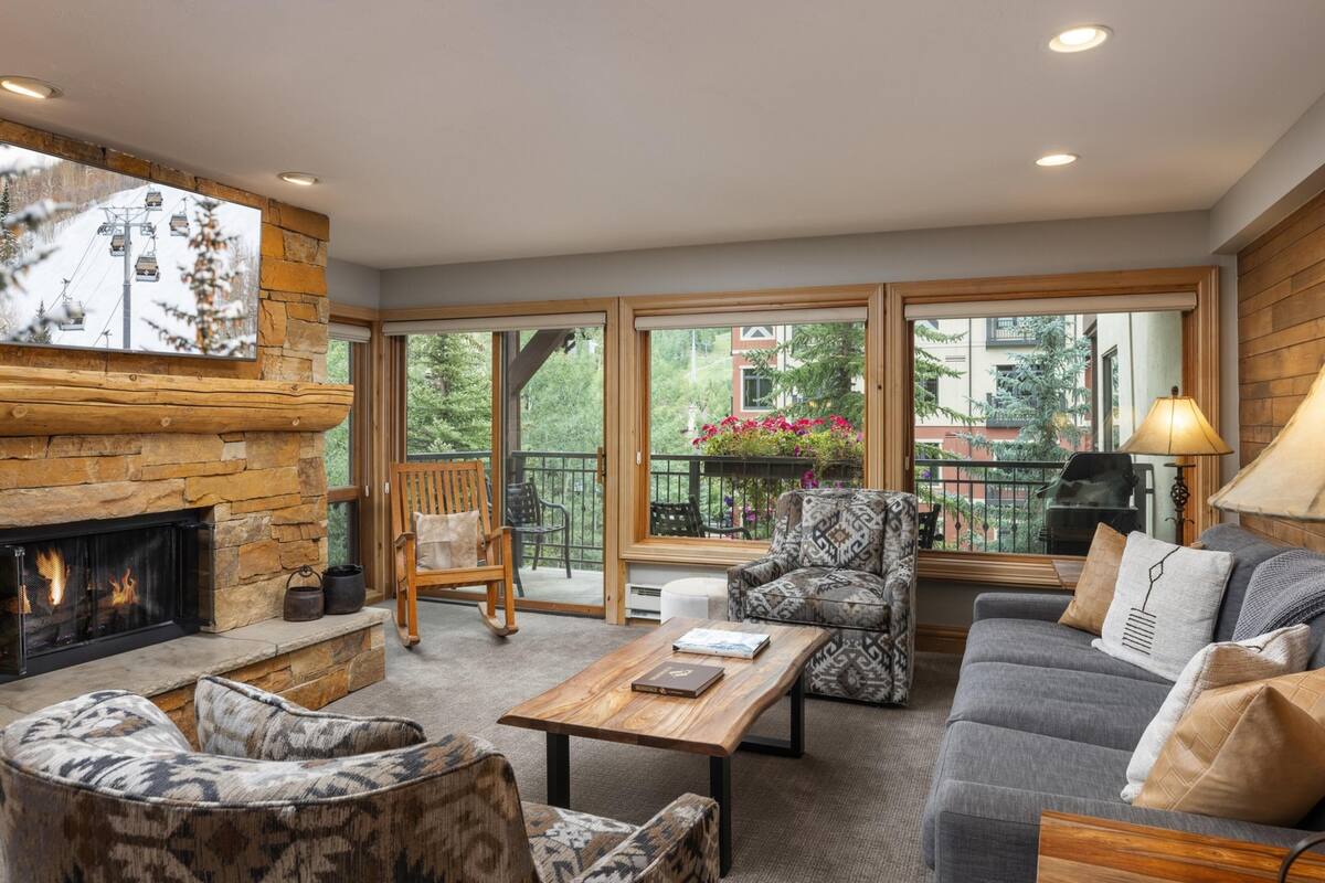 A cozy living room with a fireplace, comfortable seating, and large windows offering a view of the outdoors.