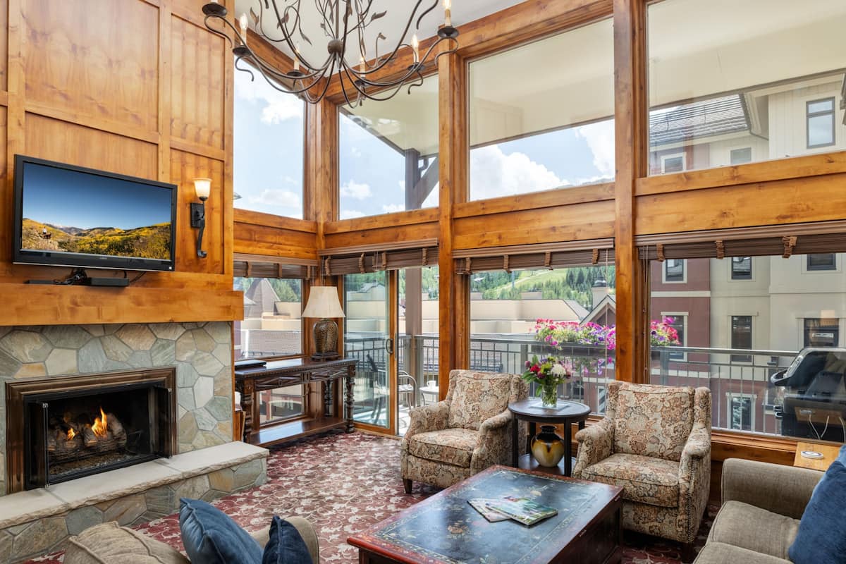 A spacious living room with a stone fireplace, comfortable seating, a television, and large windows offering a view of the surrounding area.