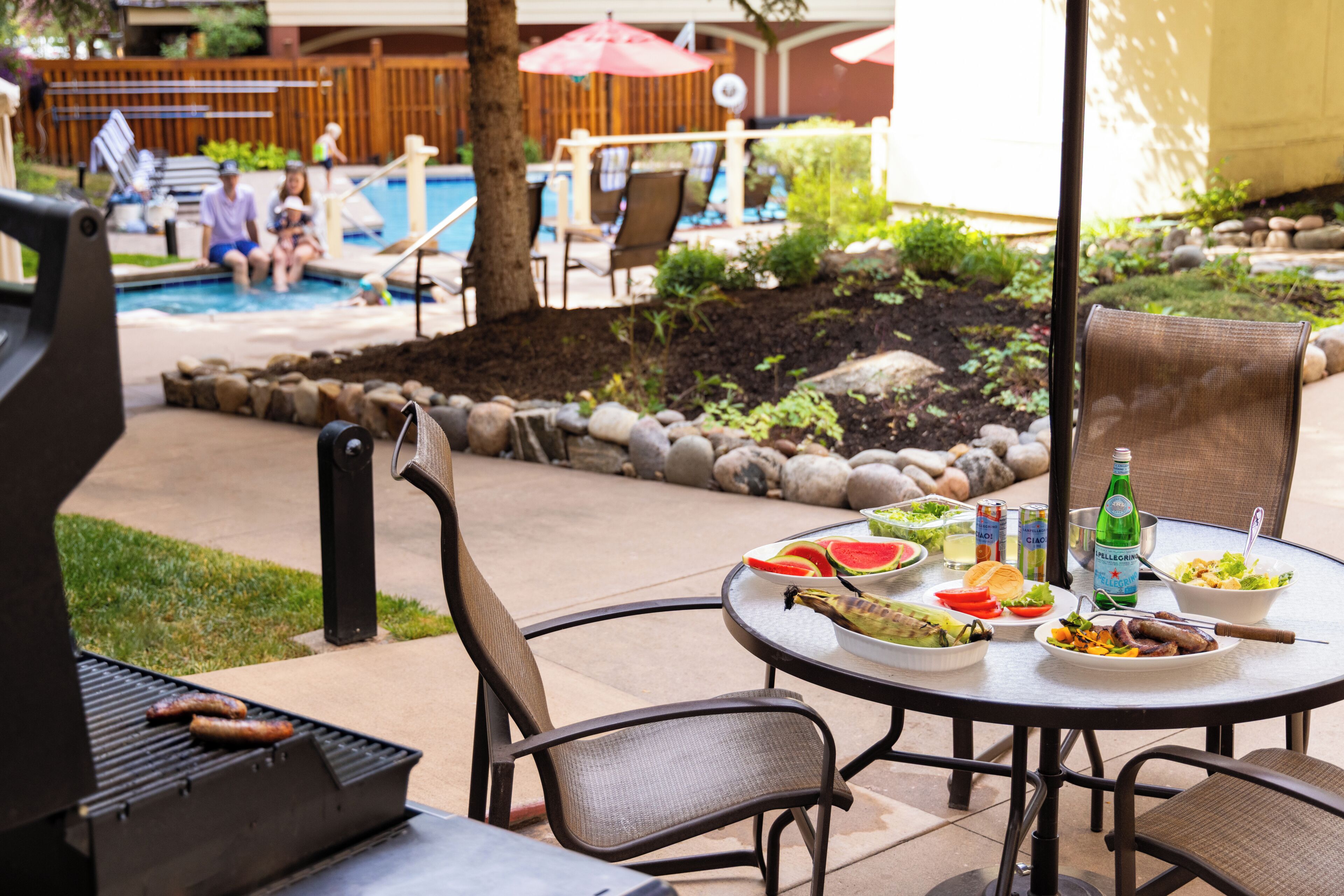 An outdoor dining area with a grill, table, chairs, and a pool in the background.