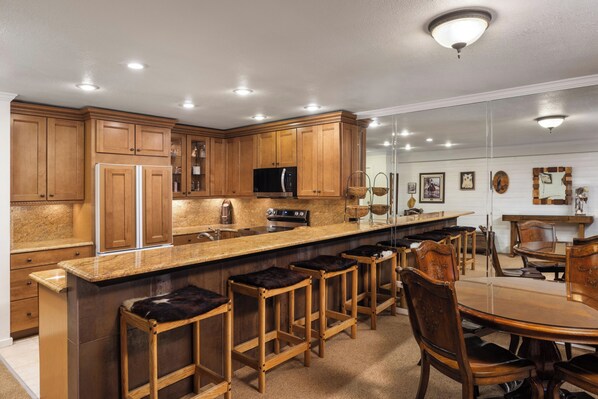 A kitchen with wooden cabinets, a central island with stools, a dining table, and a refrigerator.