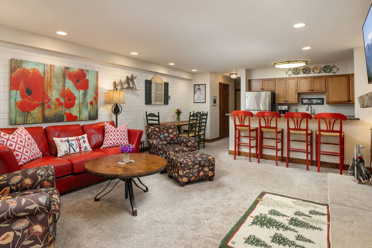 A living room with a red sofa, a patterned ottoman, and a coffee table. There is a kitchen area with wooden cabinets and a refrigerator.