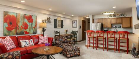 A living room with a red sofa, a patterned ottoman, and a coffee table. There is a kitchen area with wooden cabinets and a refrigerator.