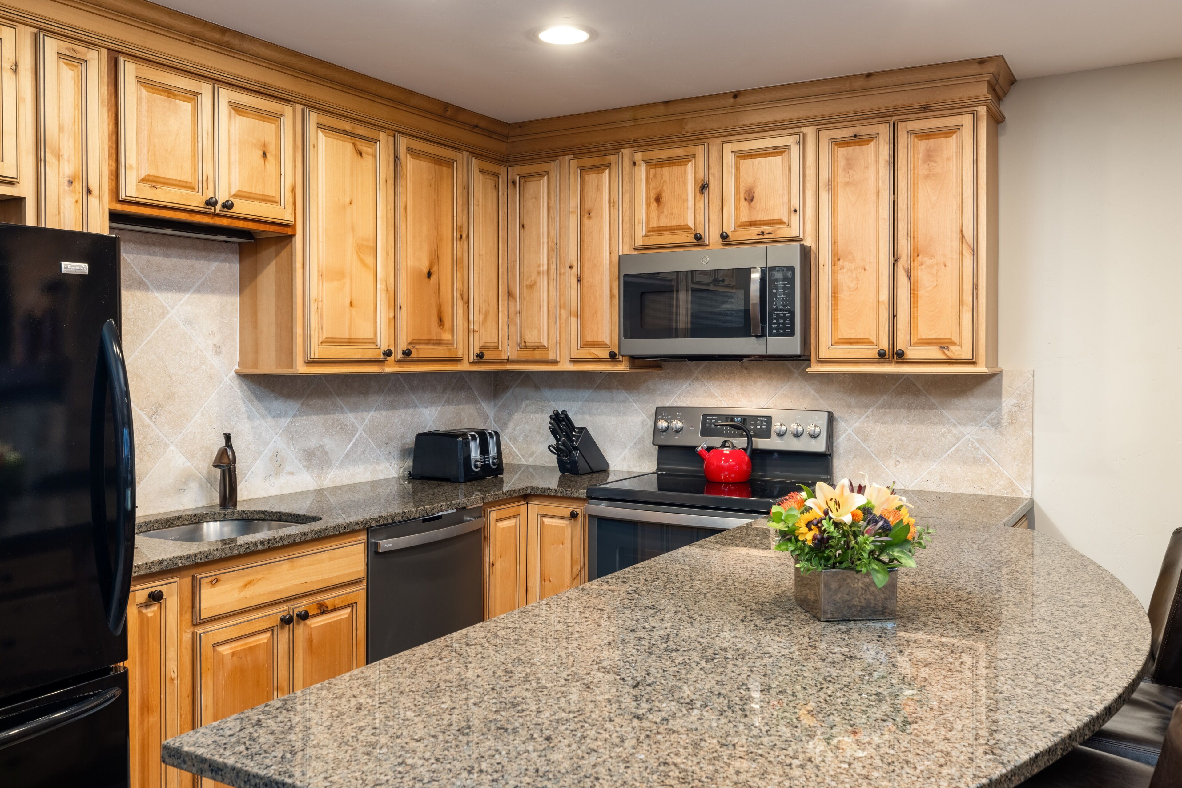 A kitchen with wooden cabinets, granite countertops, a black refrigerator, a microwave, a stove, and a flower vase.