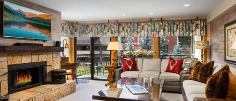 A cozy living room with a fireplace, comfortable seating, a flat-screen TV, and a balcony with a view of the mountains.