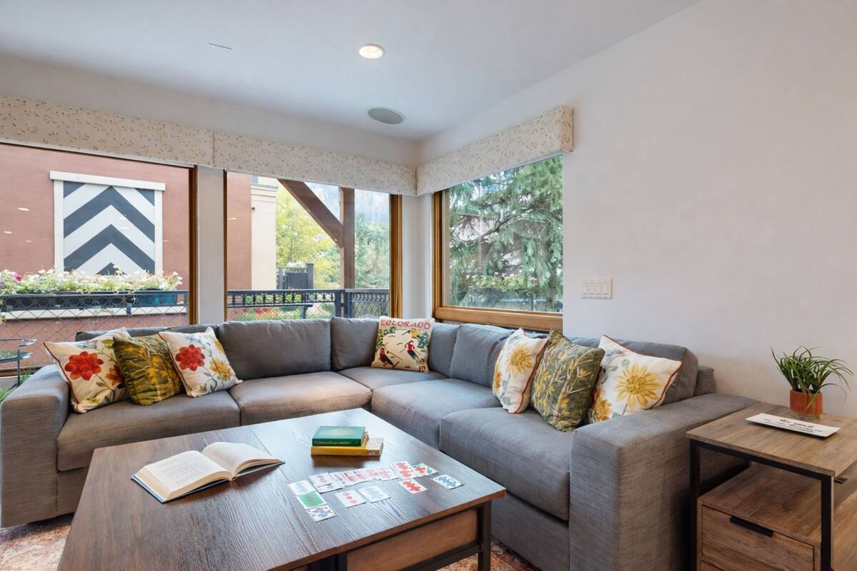 A modern living room with a grey sectional sofa, a wooden coffee table with books and playing cards, and a small wooden side table with a plant.
