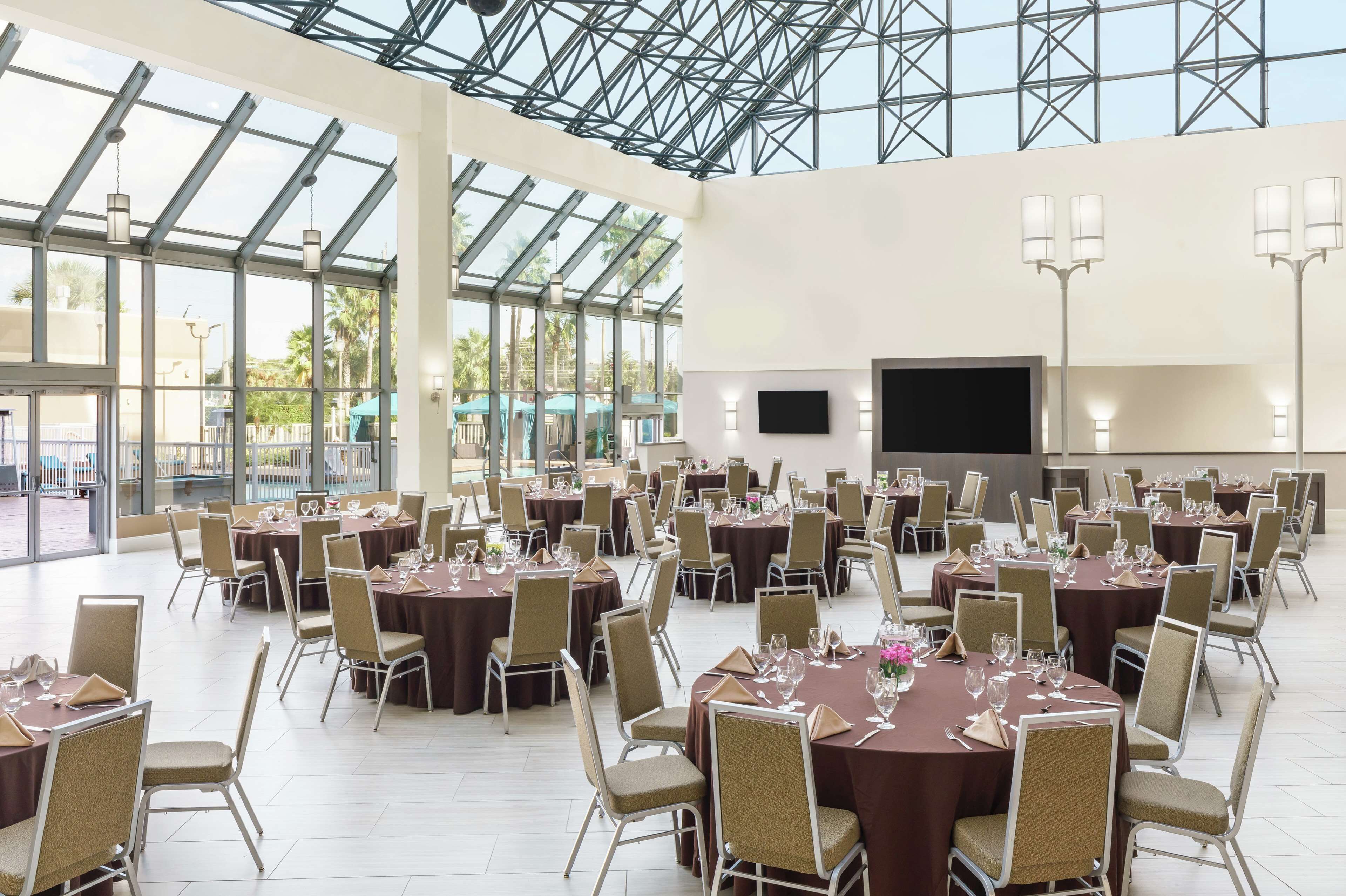 Un amplio salón de banquetes con mesas redondas y sillas, ventanas grandes y un televisor de pantalla plana.