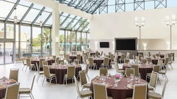 Un amplio salón de banquetes con mesas redondas y sillas, ventanas grandes y un televisor de pantalla plana.