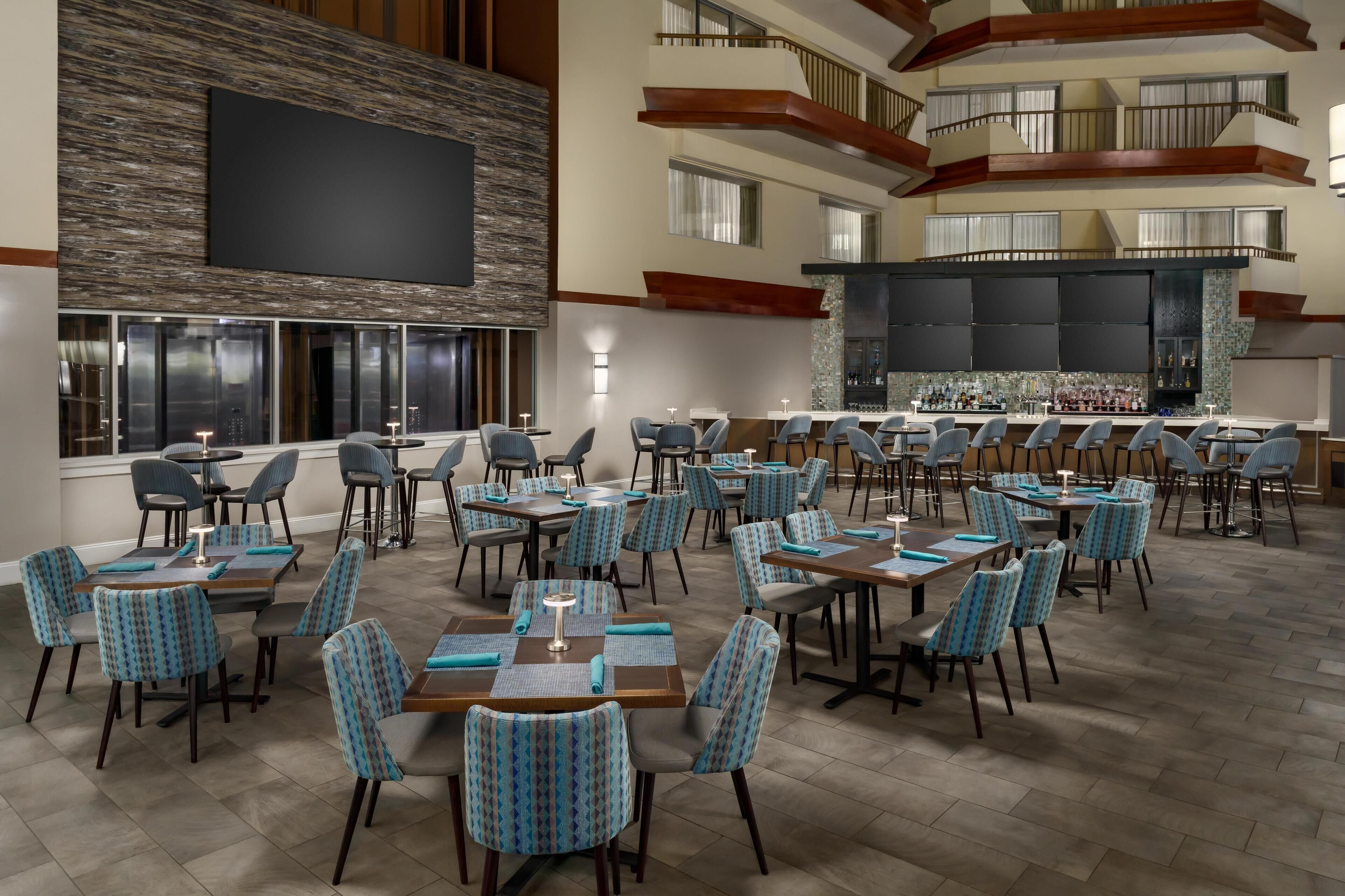 A spacious dining area with multiple tables and chairs, a bar area with a counter and stools, and a large screen on the wall.