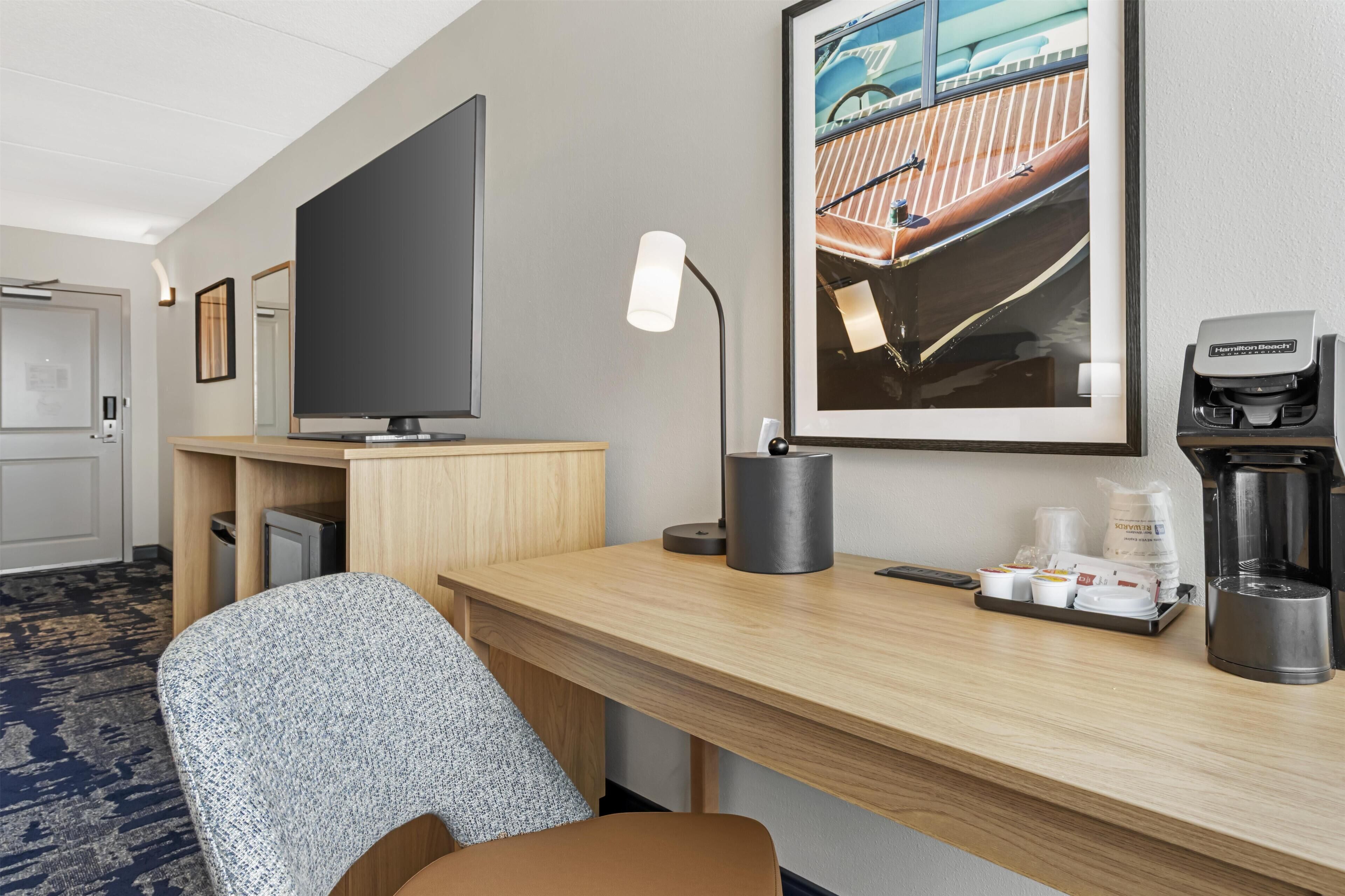 A hotel room with a desk, a chair, a television, a framed picture, a lamp, and a coffee maker.
