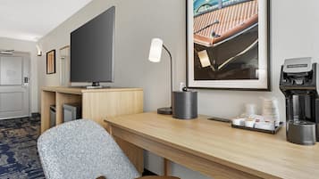 A hotel room with a desk, a chair, a television, a framed picture, a lamp, and a coffee maker.